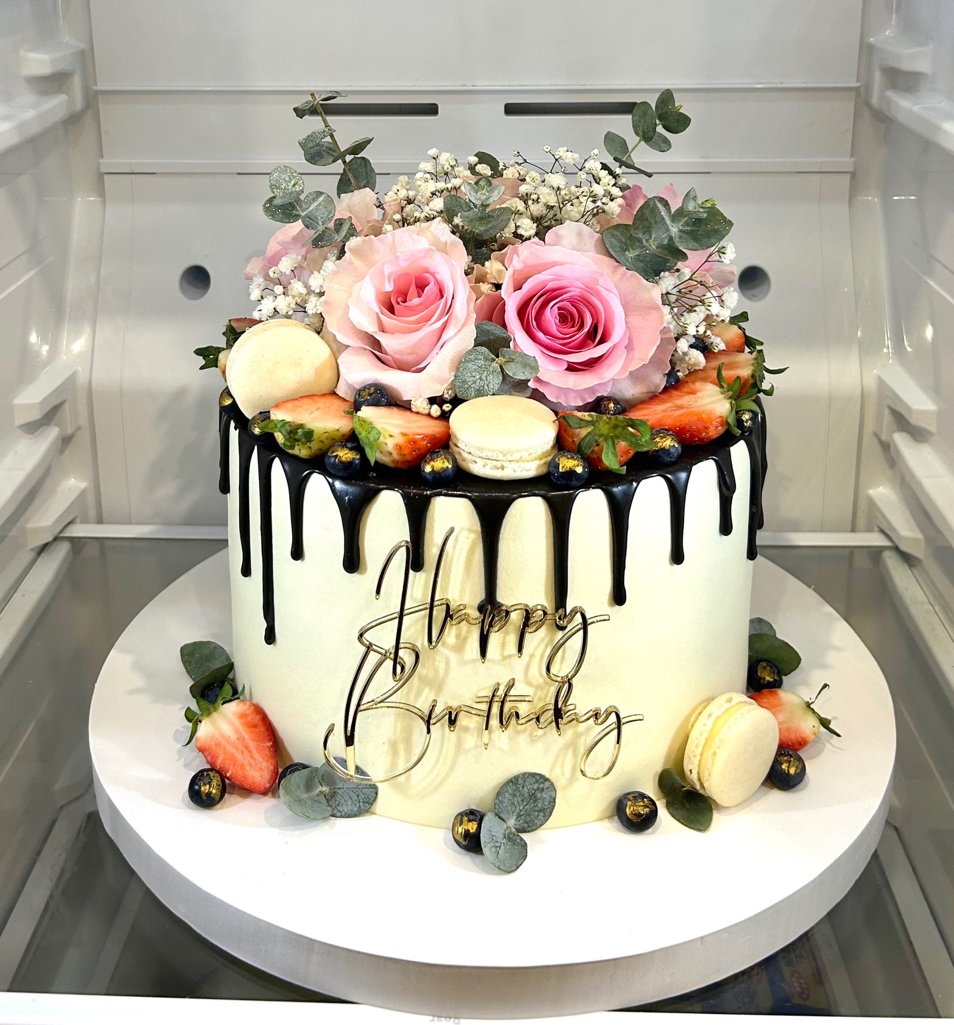 Tort urodzinowy z kremem, czarną polewą, makaronikami, truskawkami, borówkami i różami, umieszczony na białym podeście w chłodnym otoczeniu.