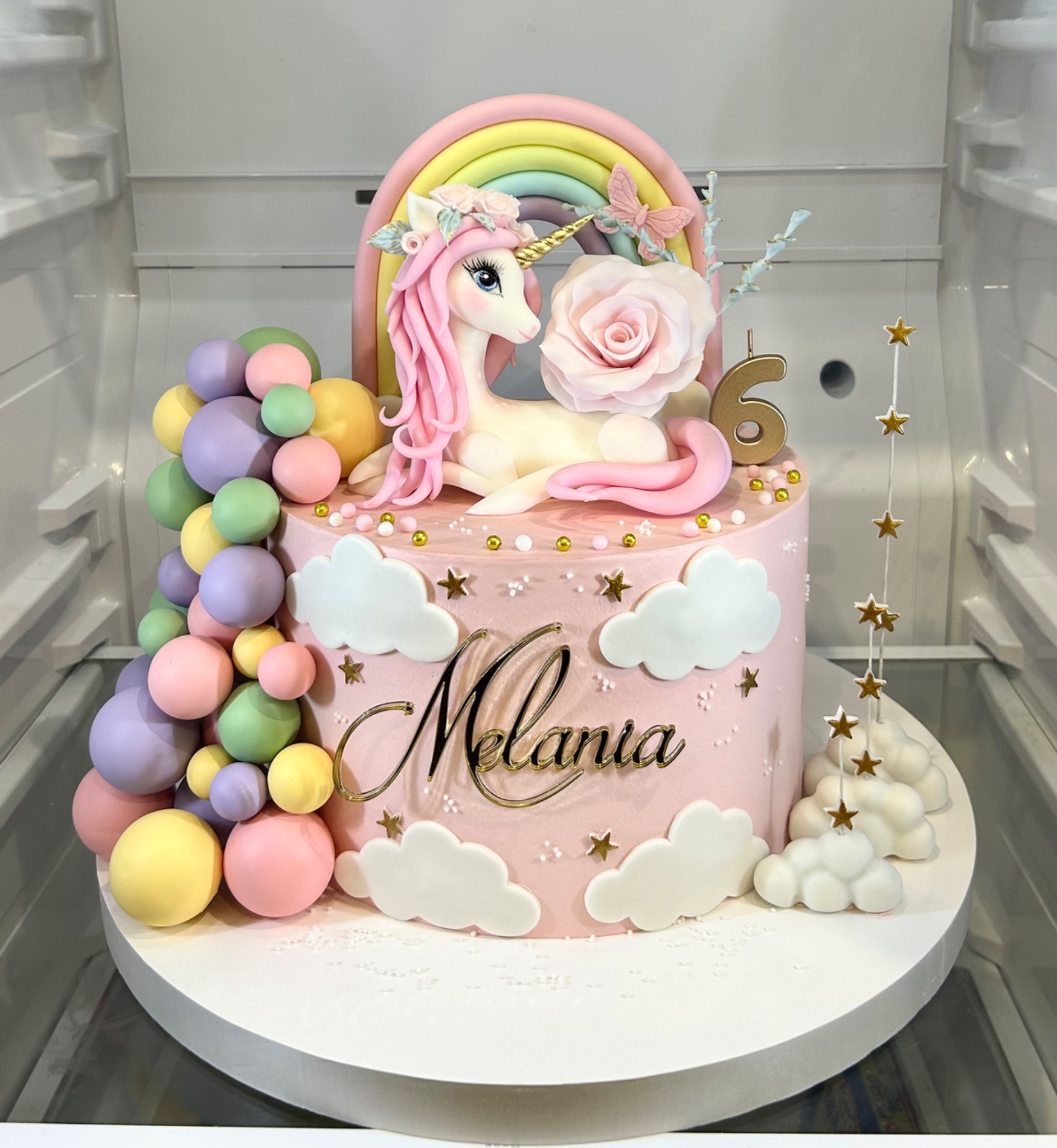 Różowy tort urodzinowy z jednorożcem, tęczą, balonami i imieniem 'Melania', dekorowany chmurkami i gwiazdkami, umieszczony w chłodziarce.