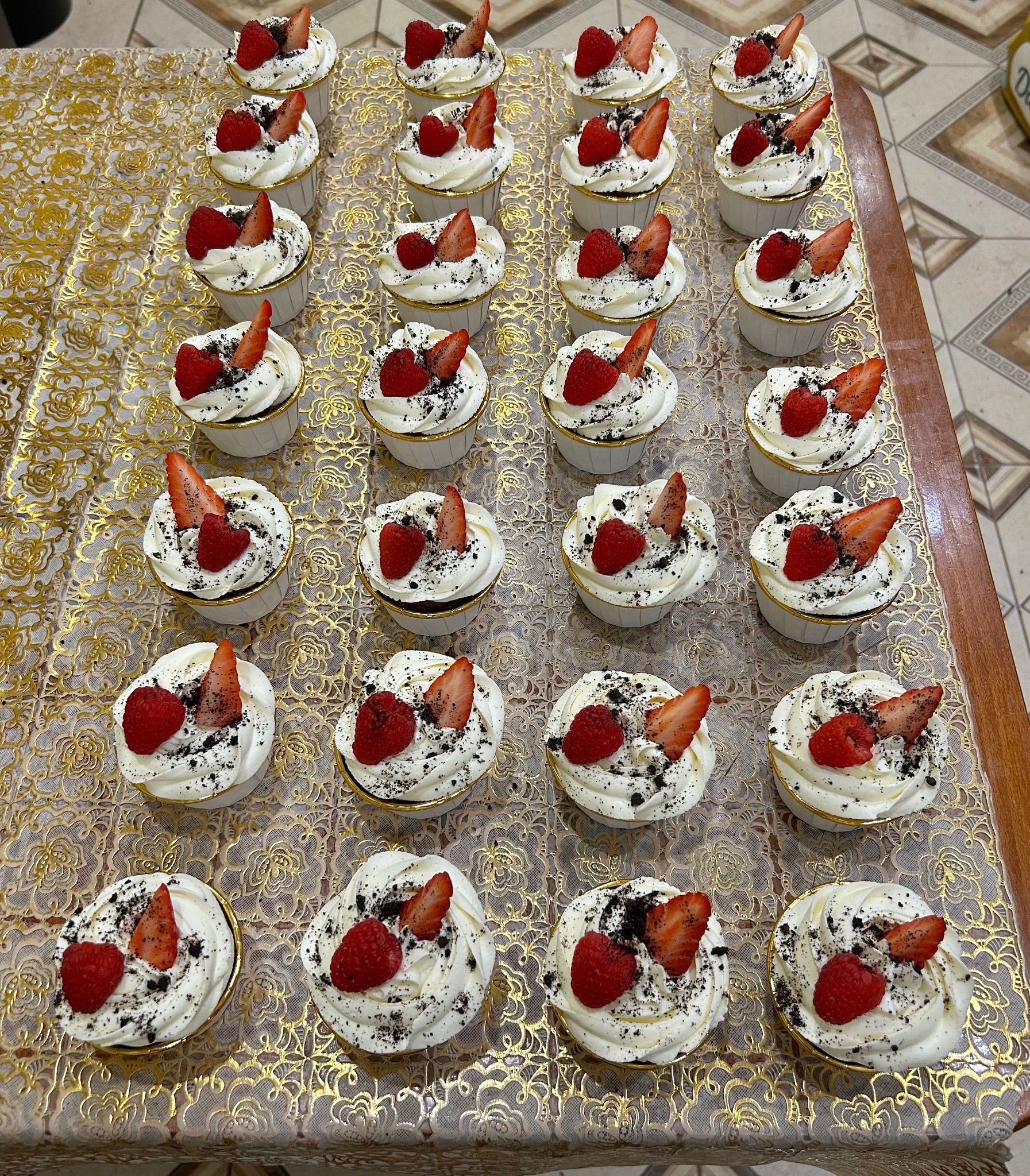 Rząd babeczek z kremem, posypką i owocami (maliny, truskawki) na złotej, wzorzystej serwetce. Widok z góry, prezentacja wyrobów.