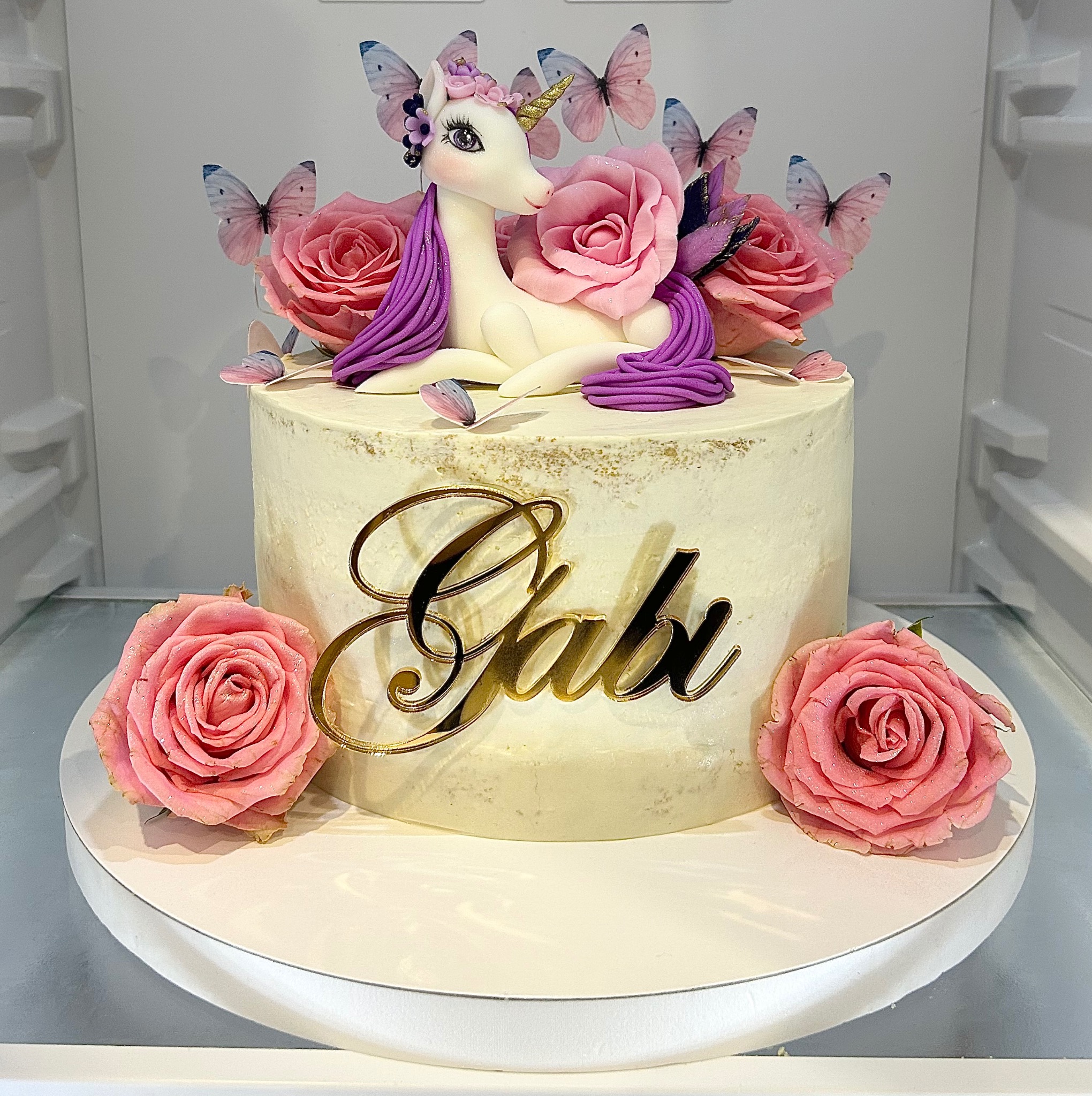 Tort z jednorożcem, różami i motylami, z napisem 'Gabi' w złotym kolorze. Kremowy tort na białym podeście, w tle biała lodówka. Dekoracja urodzinowa.