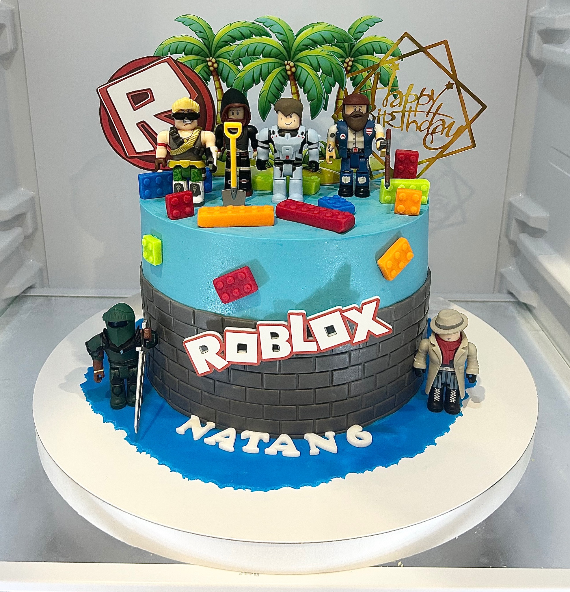 Dwupoziomowy tort urodzinowy w stylu Roblox, z figurkami postaci, palmami i napisem 'Happy Birthday'. Dekoracja z klocków i imieniem 'Natan6'.