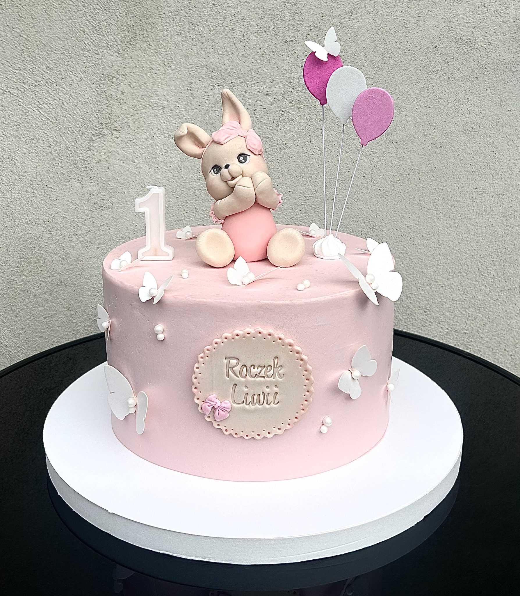 Tort na roczek z różowym kremem, figurką króliczka, motylami i balonami. Napis 'Roczek Liwii'. Dekoracja na stół urodzinowy, wykonana z dbałością o detale.