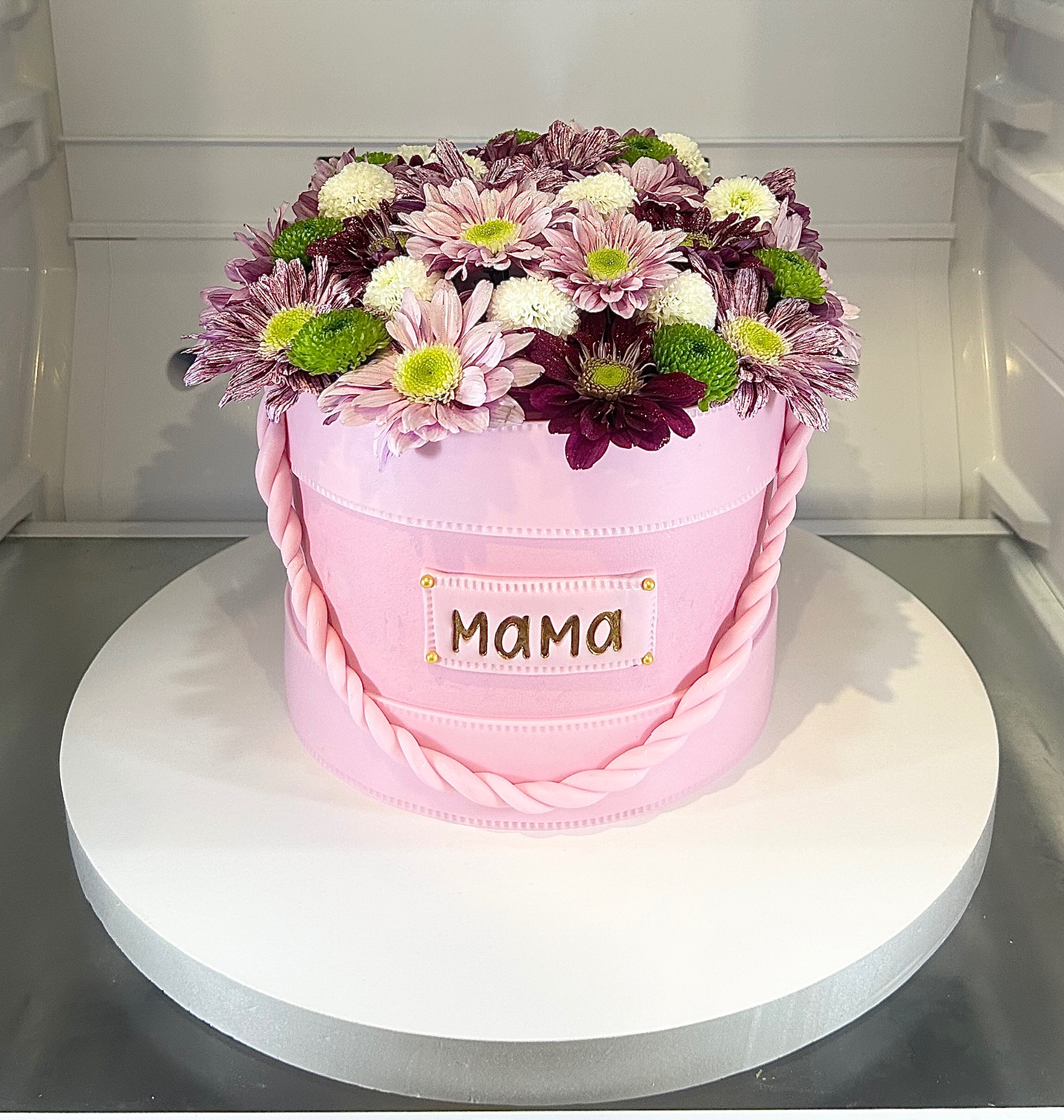 Różowy tort w kształcie pudełka z kwiatami, dekorowany napisem 'Mama' w złotym kolorze, umieszczony na białym obrotowym talerzu w chłodnym otoczeniu.