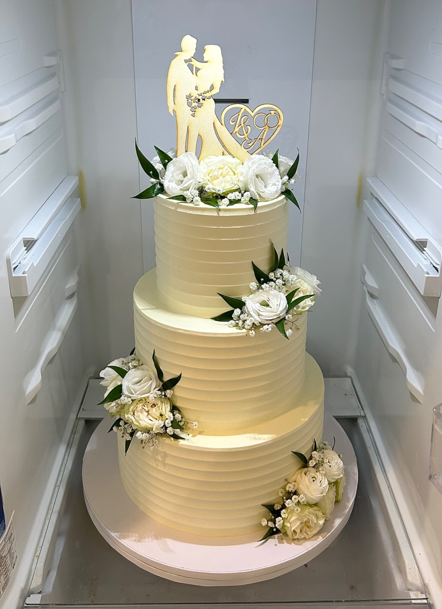 Trzypiętrowy tort weselny w chłodnym odcieniu bieli, zdobiony świeżymi białymi kwiatami i drewnianą ozdobą z sylwetkami pary młodej, umieszczony w chłodziarce.