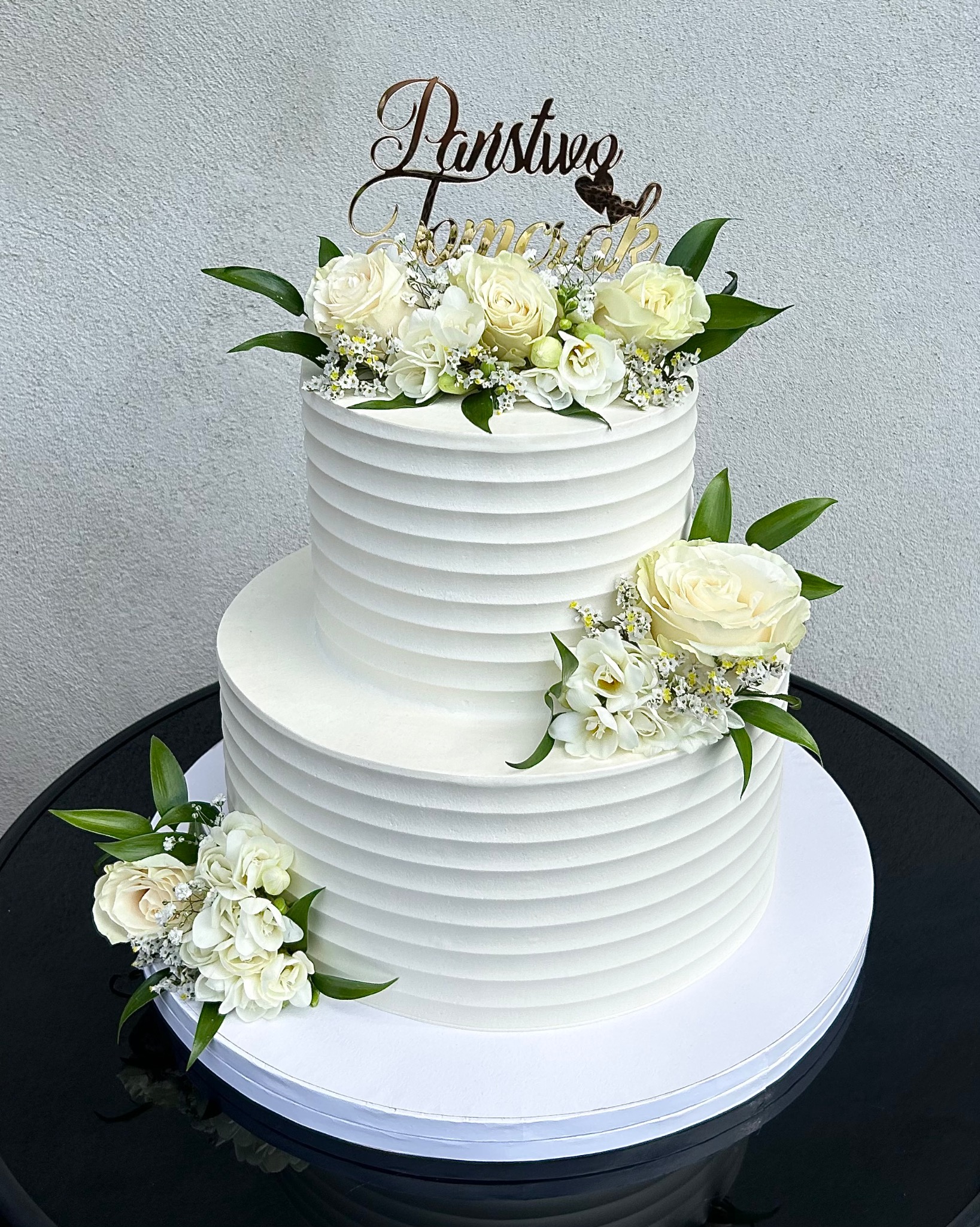 Dwupoziomowy tort weselny z białym kremem, zdobiony białymi różami i topperem 'Państwo Młodzi', na czarnym stole. Minimalistyczny design, elegancki wygląd.