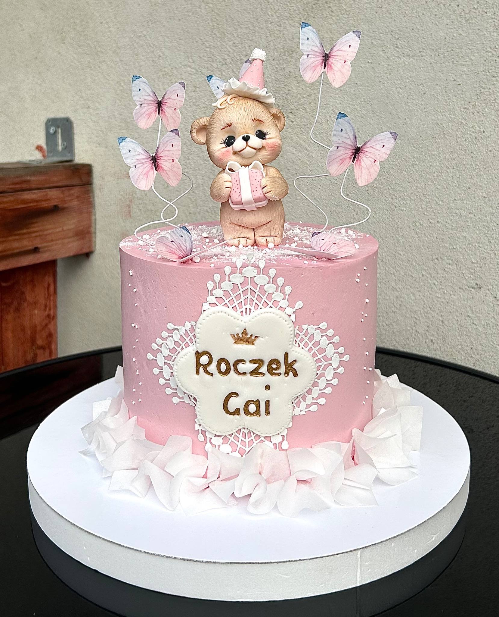Różowy tort na roczek z misiem w czapeczce i motylami, dekorowany białymi koronkami i napisem 'Roczek Gai'. Uroczy i słodki wypiek na pierwsze urodziny.