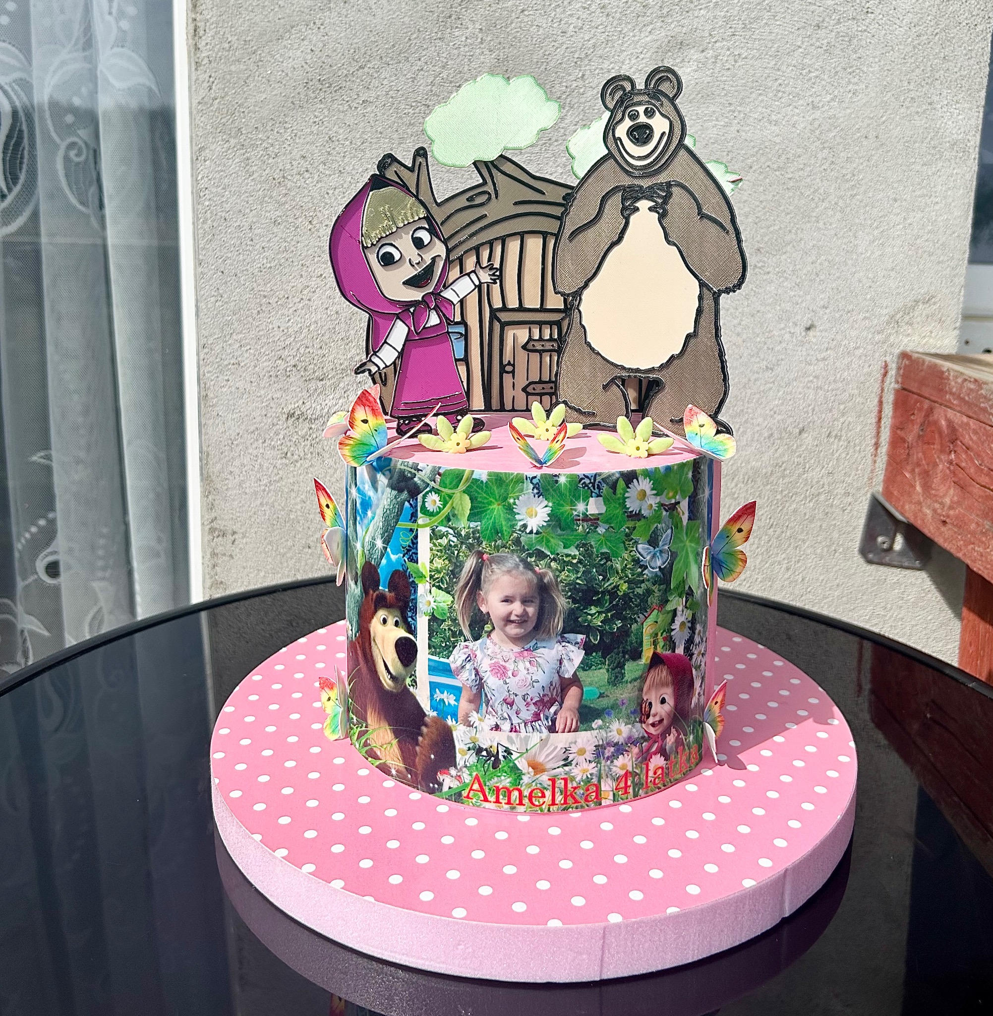 Tort urodzinowy z motywem Masha i Niedźwiedź, ozdobiony zdjęciem dziecka i motylami, na różowym podkładzie w białe kropki, stojący na czarnym stole.