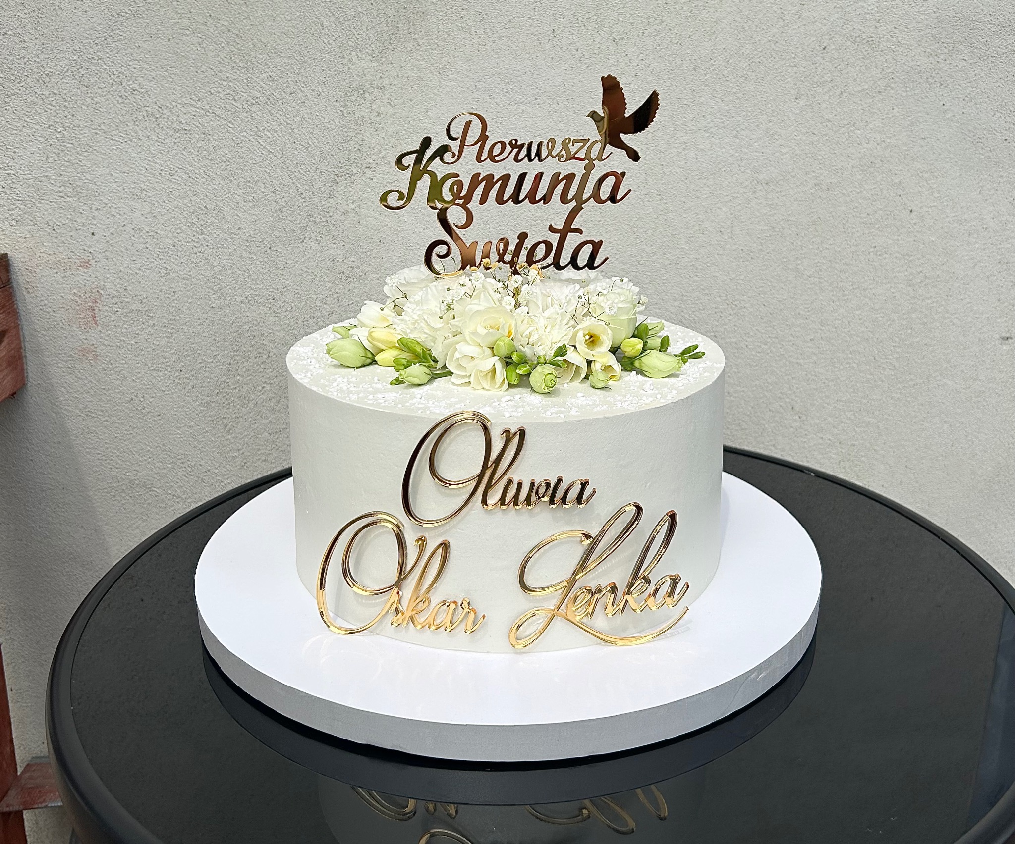Tort komunijny z białym kremem, złotymi napisami imion i topperem 'Pierwsza Komunia Święta' z gołębiem, udekorowany białymi kwiatami na czarnym stole.
