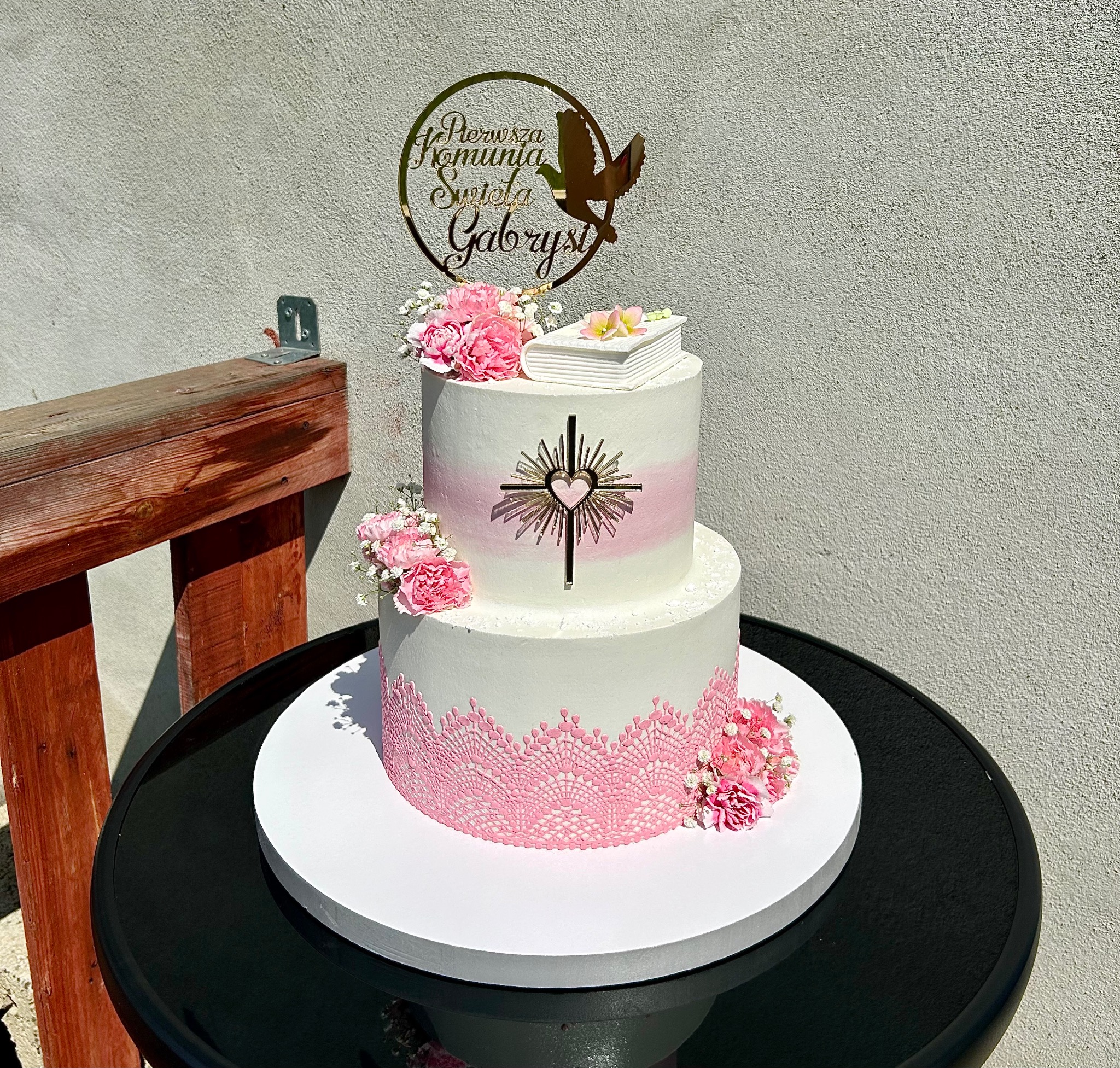 Dwupoziomowy tort komunijny w bieli i różu, zdobiony kwiatami i złotym topperem z gołębiem, na czarnym stoliku z drewnianym elementem w tle.