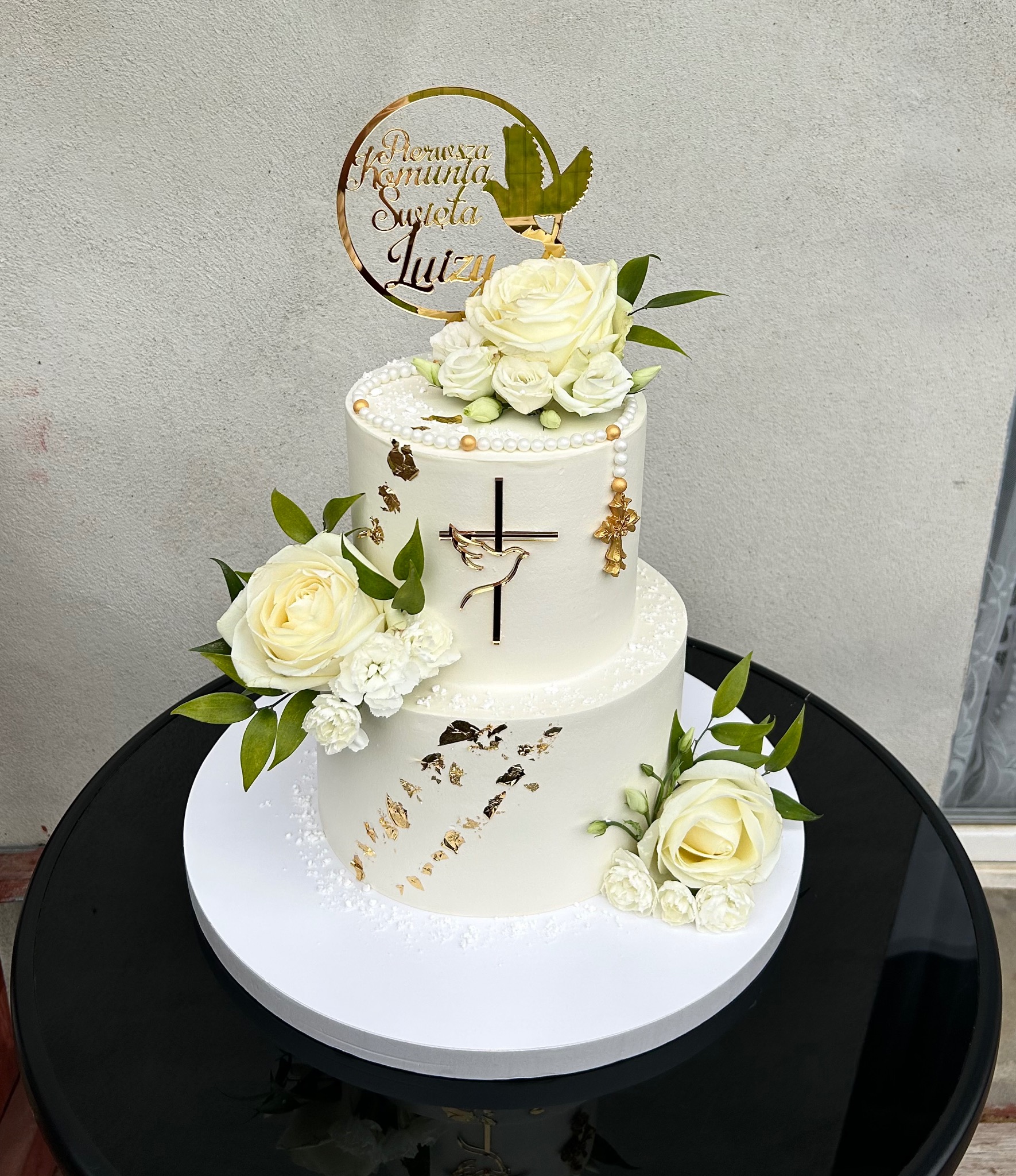 Dwupoziomowy tort komunijny w bieli, zdobiony złotymi akcentami, białymi różami i topperem z napisem 'Pierwsza Komunia Święta Luizy'. Elegancki i delikatny.