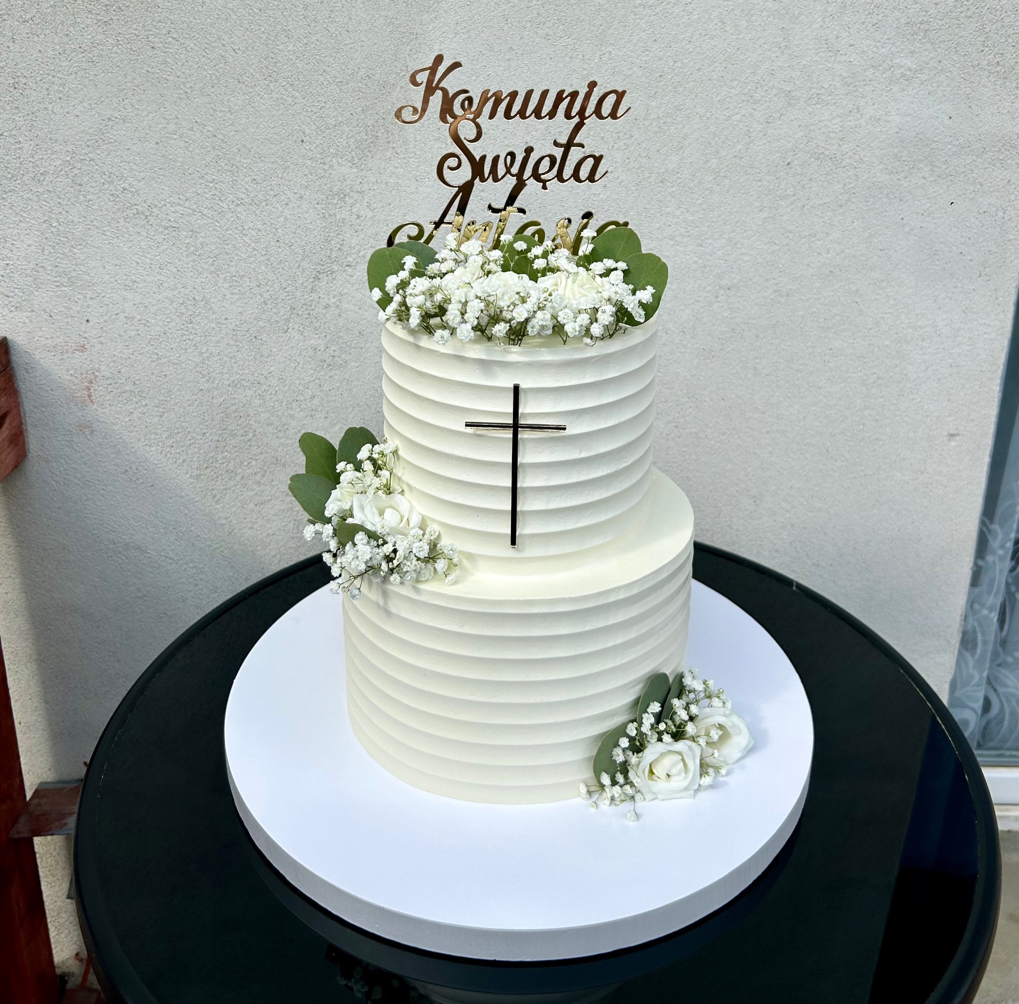 Dwupiętrowy tort komunijny z kremowym wykończeniem, dekorowany białymi kwiatami i złotym topperem z napisem 'Komunia Święta Antoniego', na czarnym stole.