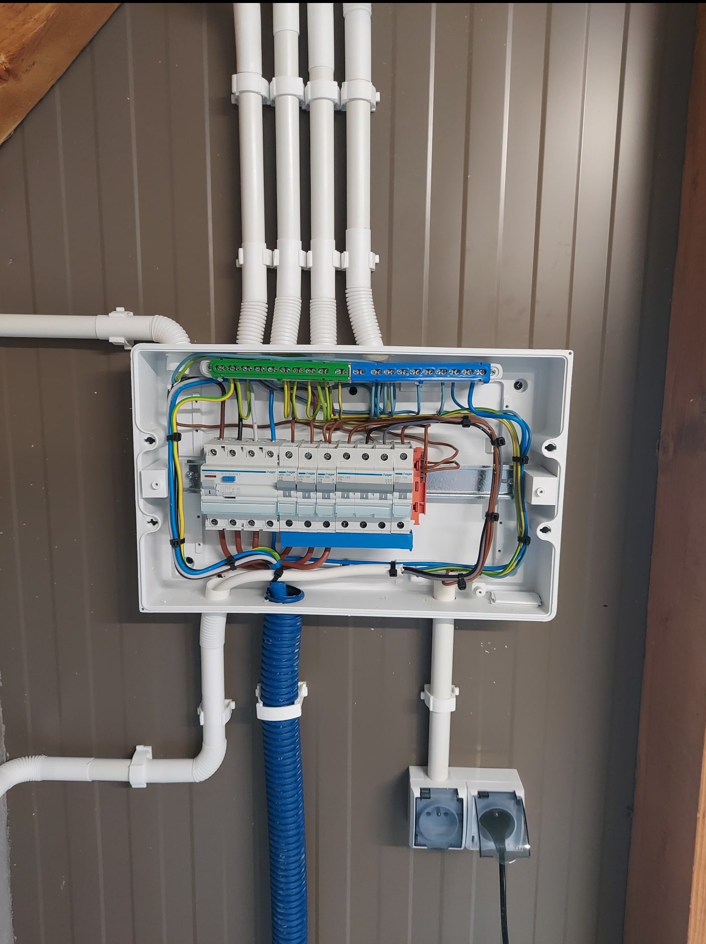 Rozdzielnica elektryczna w białej obudowie z widocznymi przewodami, bezpiecznikami i listwami zaciskowymi. Białe rury instalacyjne prowadzą do góry i do dołu. Gniazdko z wtyczką.