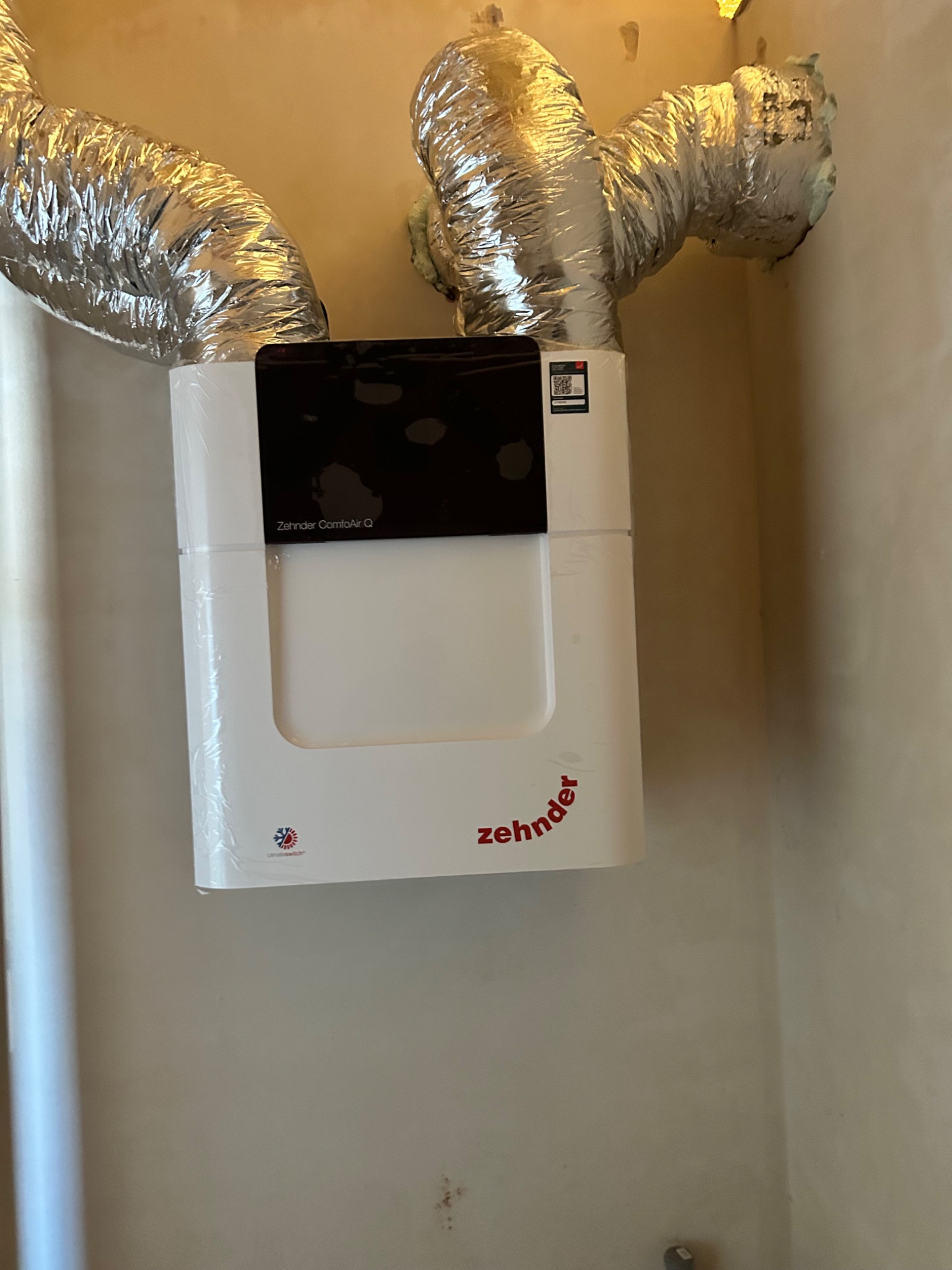 Biała jednostka rekuperacyjna Zehnder ComfoAir Q z podłączonymi, srebrnymi przewodami wentylacyjnymi na tle beżowej ściany. Widoczne ślady po montażu.
