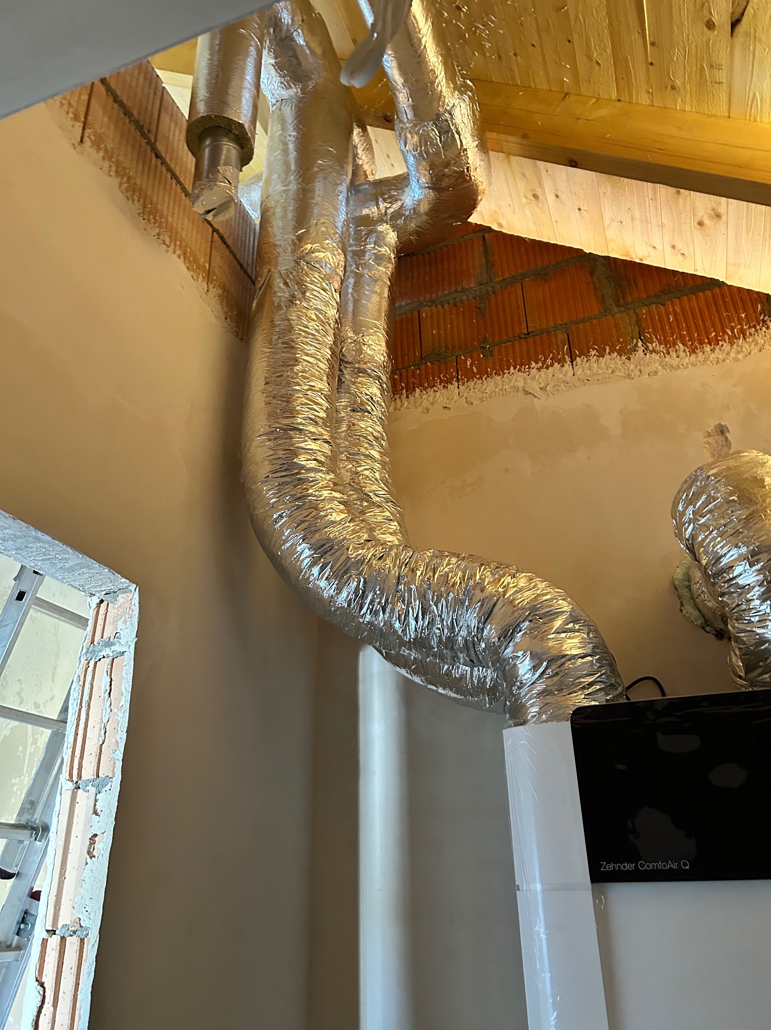 Instalacja rekuperacji z widocznymi przewodami wentylacyjnymi w srebrnej izolacji, zamontowanymi pod drewnianym stropem. Widoczny fragment urządzenia Zehnder ComfoAir Q.