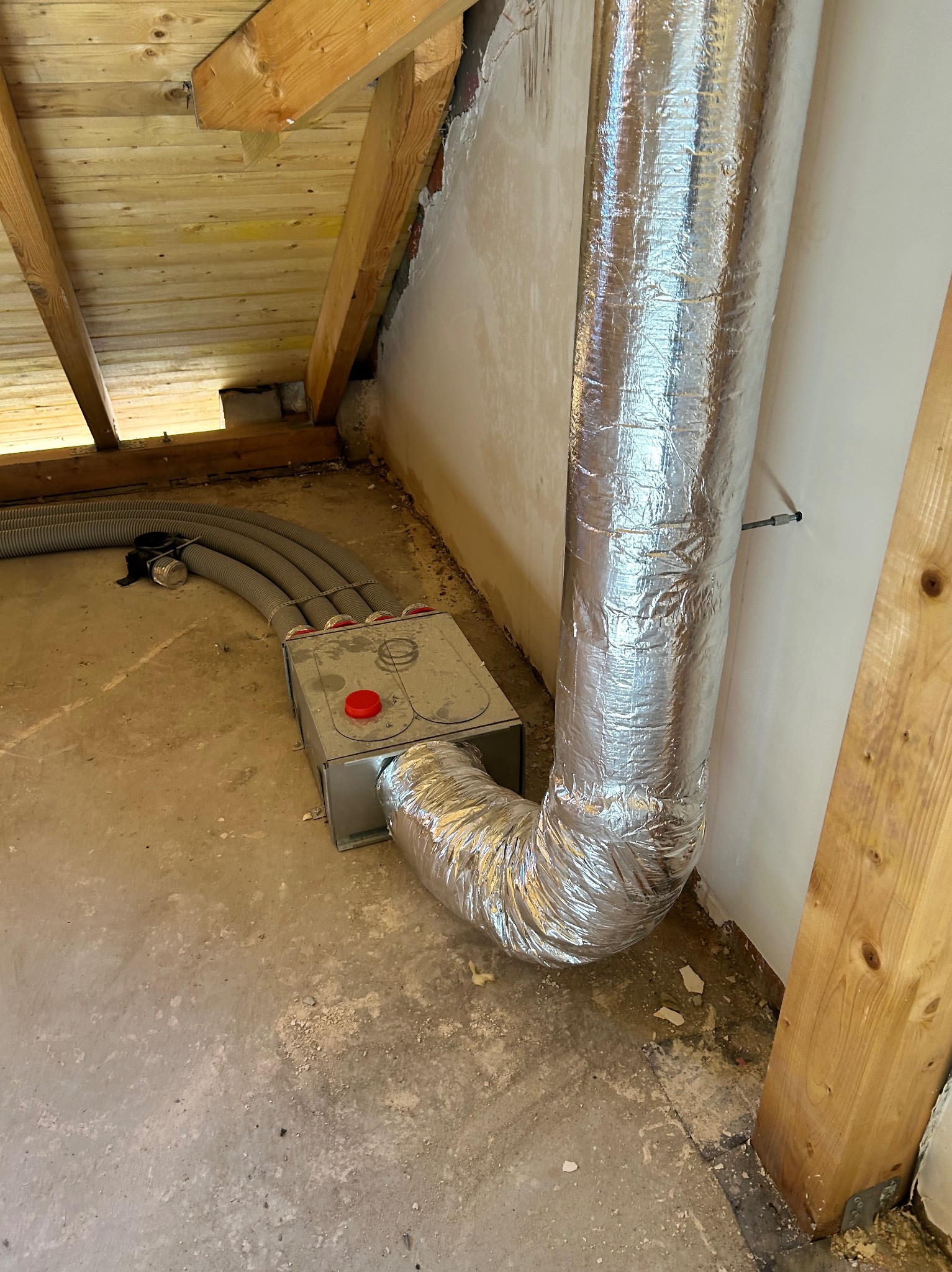 Szara skrzynka z czerwonym przyciskiem i srebrna rura wentylacyjna w surowym wnętrzu poddasza z drewnianą konstrukcją dachu. Widoczne szare, elastyczne przewody.