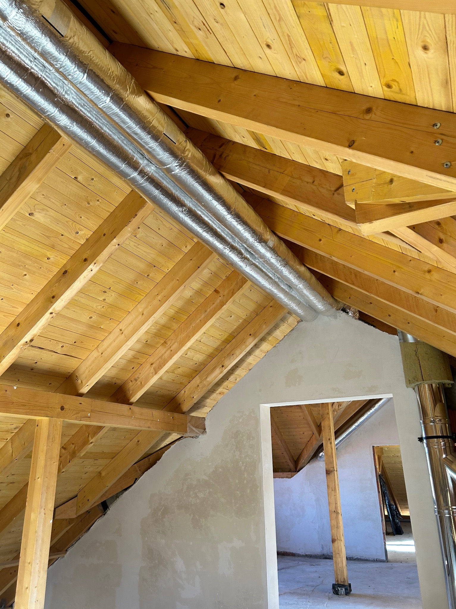 Dwa srebrne przewody wentylacyjne zamontowane pod drewnianym dachem na poddaszu. Widoczne elementy konstrukcyjne dachu i fragment ściany z otworem drzwiowym.