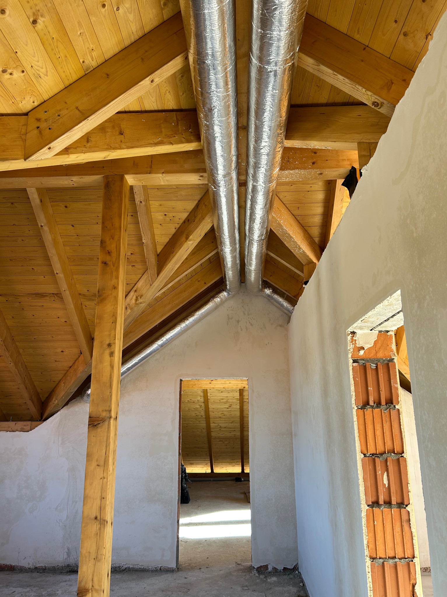 Dwa srebrne przewody wentylacyjne pod drewnianym stropem poddasza w surowym stanie. Widoczne elementy konstrukcyjne i fragmenty ścian z cegły.