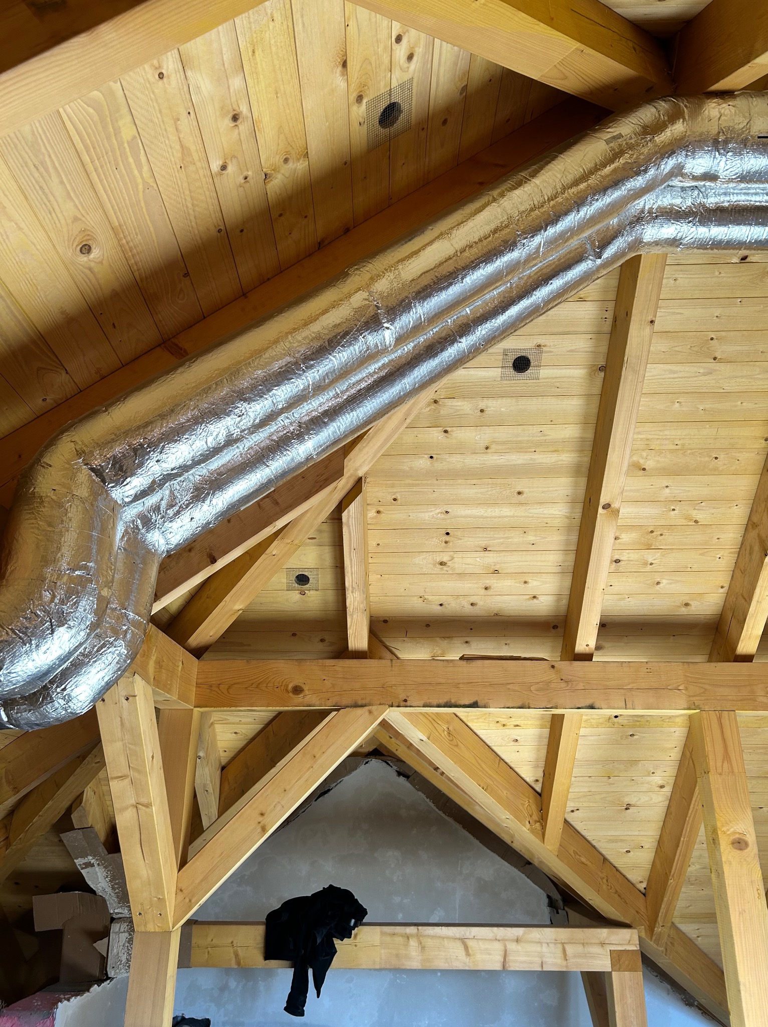 Izolowany przewód wentylacyjny z folii aluminiowej zamontowany pod drewnianym dachem z boazerii. Widoczne elementy konstrukcji dachu i fragment ściany w tle.