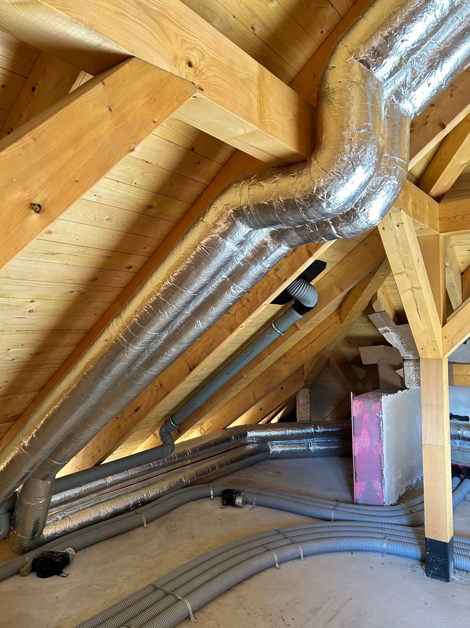 Montaż instalacji wentylacyjnej na poddaszu z widocznymi rurami wentylacyjnymi i drewnianą konstrukcją dachu. Rury w izolacji aluminiowej i szare rury karbowane.
