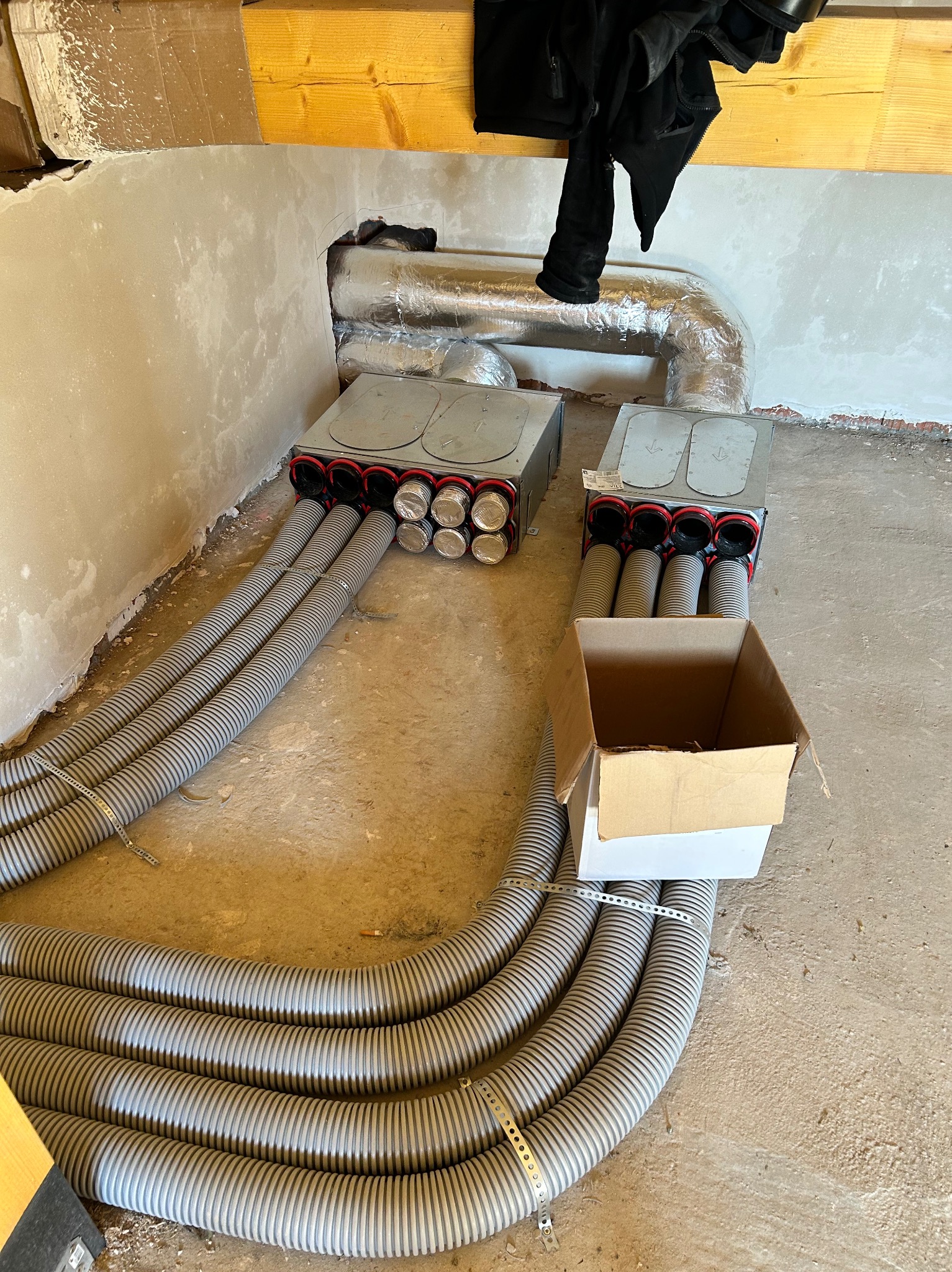 Szare, elastyczne rury wentylacyjne podłączone do metalowych skrzynek na podłodze w surowym wnętrzu. Widoczne elementy instalacji i kartonowe pudełko.