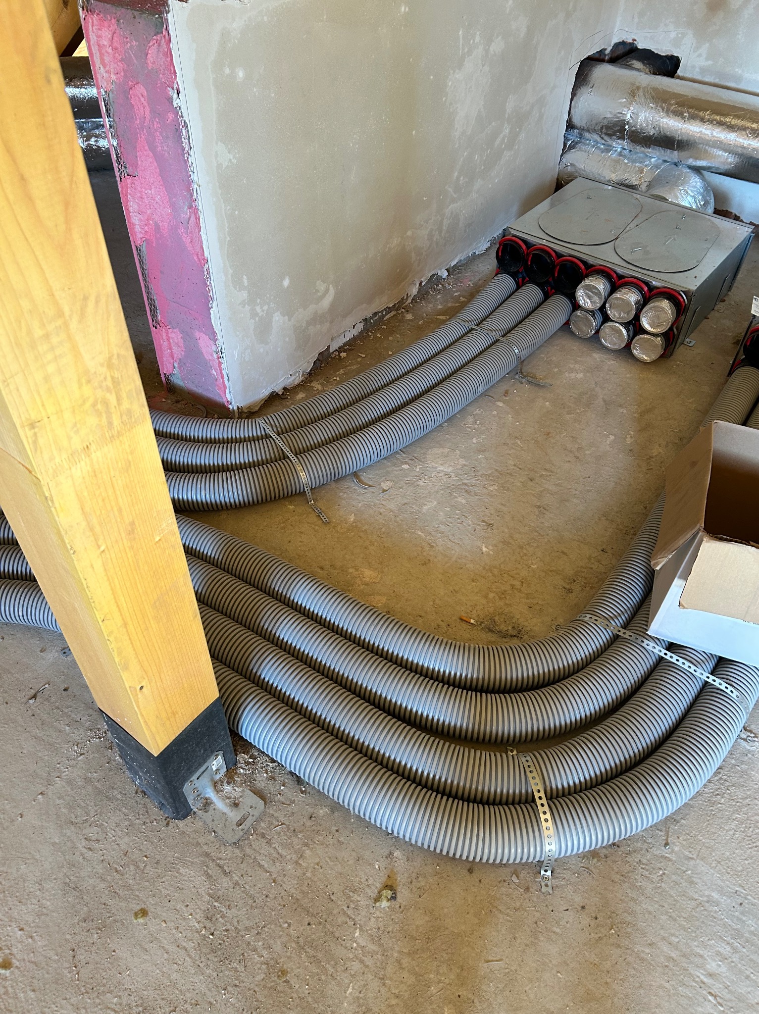 Szare, karbowane rury wentylacyjne prowadzą do metalowej skrzynki z czerwonymi obwódkami w surowym wnętrzu. Widoczny fragment drewnianej konstrukcji i surowy beton.