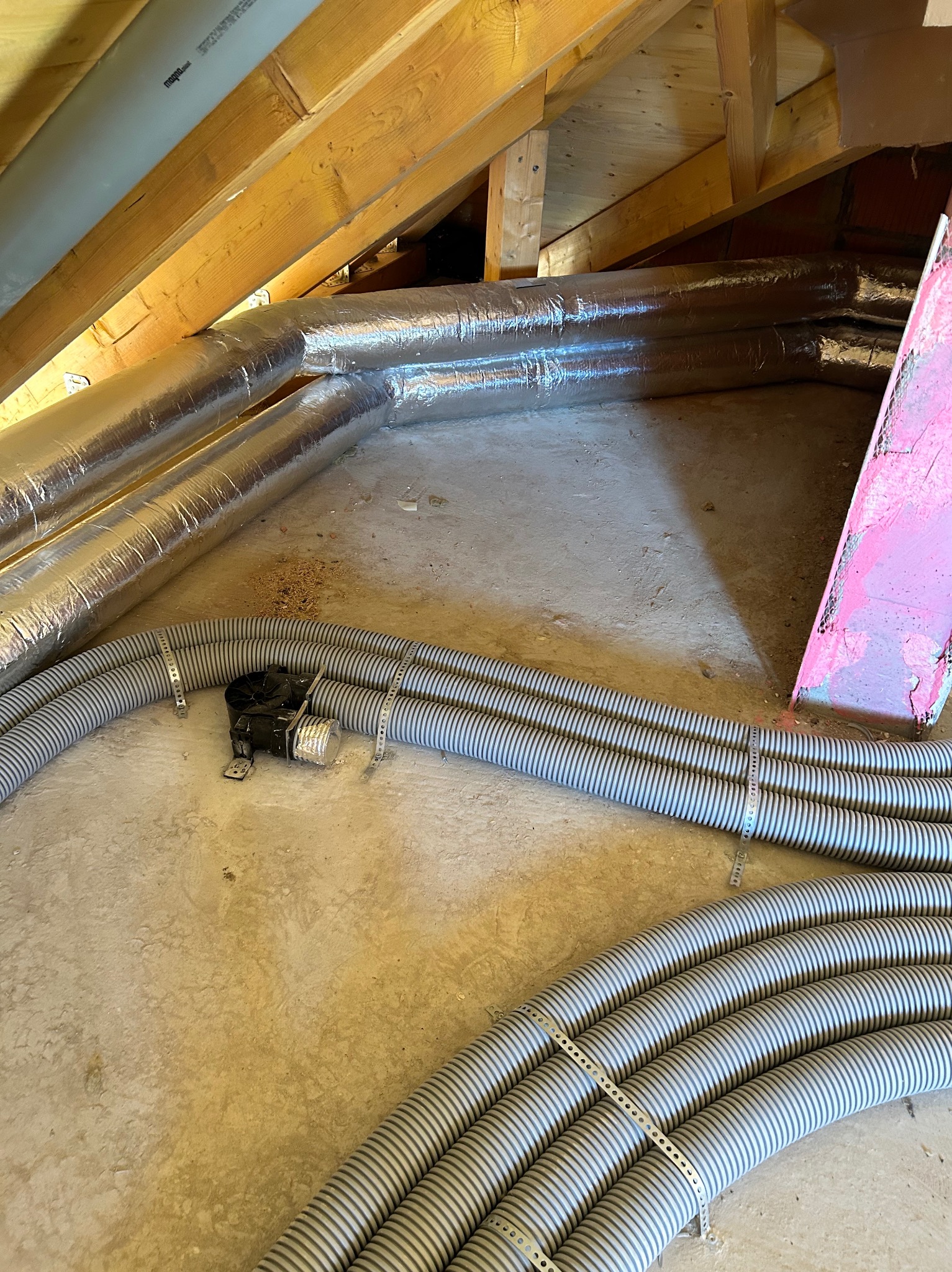 Szare, elastyczne rury wentylacyjne z metalowymi obejmami na poddaszu, obok izolowanych przewodów i drewnianej konstrukcji dachu. Widoczny fragment instalacji.