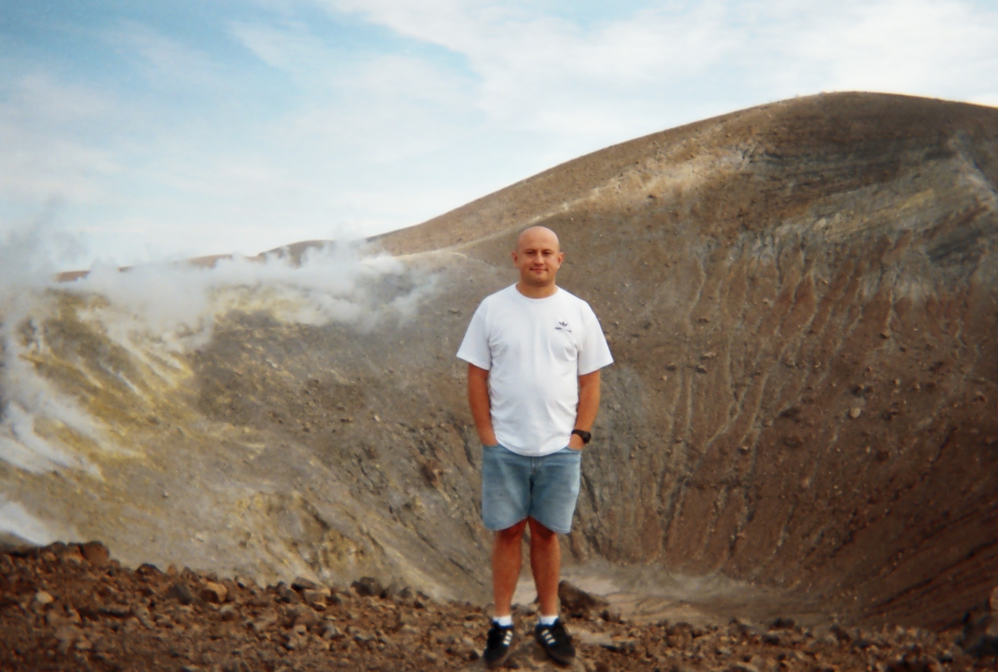 Mężczyzna w białej koszulce i dżinsowych szortach stoi na tle krateru wulkanu z wydobywającą się parą. Ujęcie z dołu, widoczne skały i niebo.