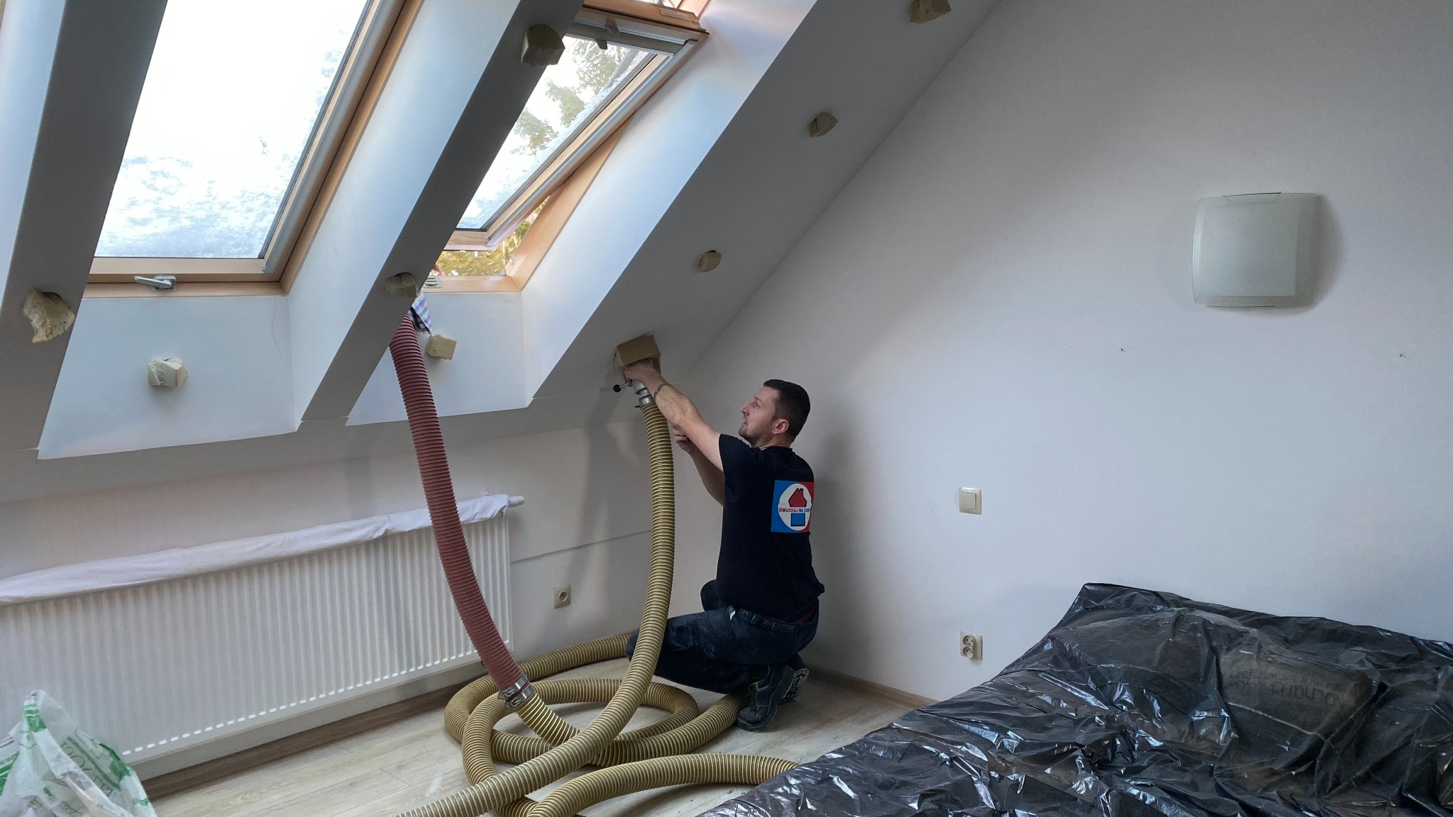 Pracownik w trakcie wdmuchiwania izolacji w poddasze z oknami dachowymi, używając węża i specjalistycznego sprzętu. Zabezpieczone meble folią ochronną.