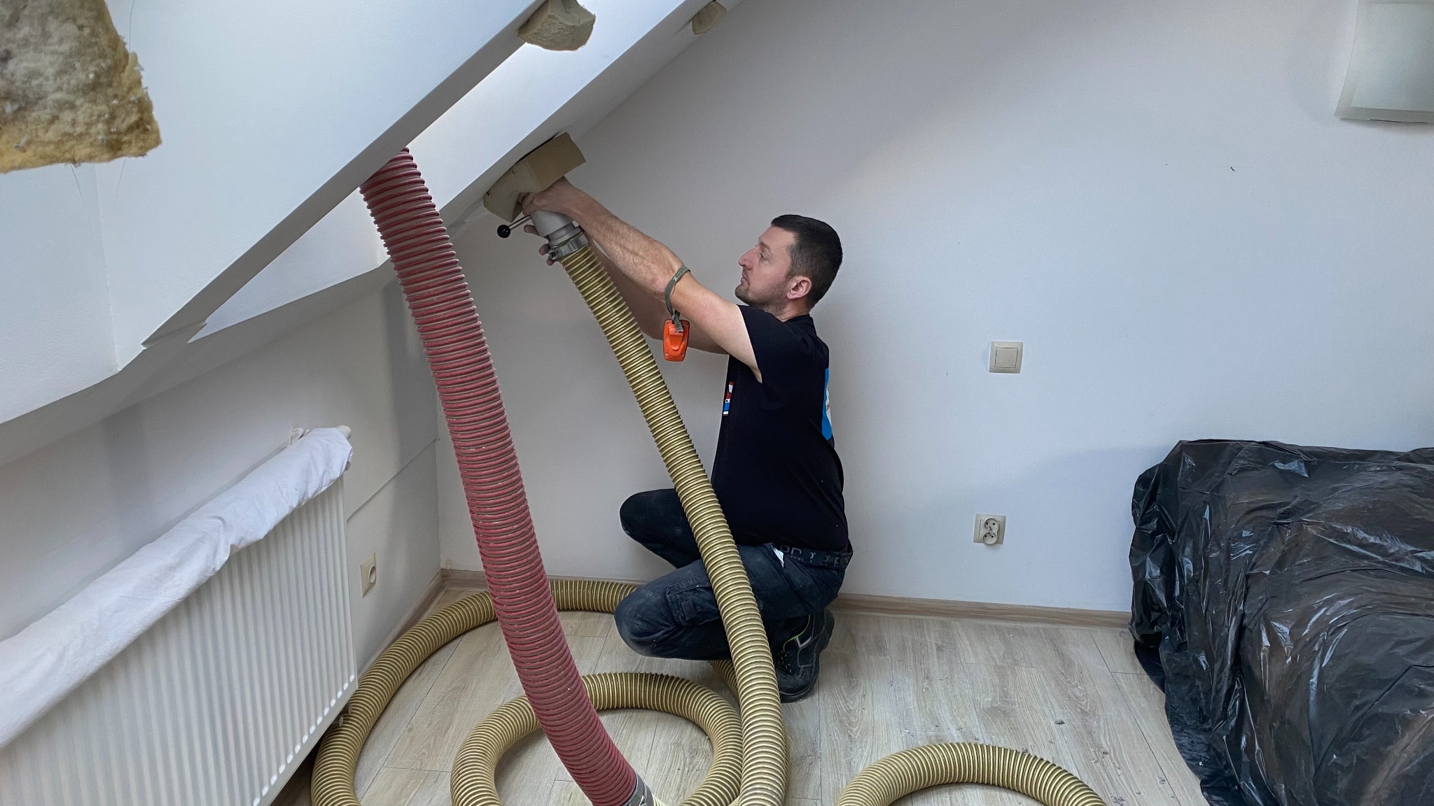 Pracownik w trakcie wdmuchiwania izolacji do poddasza, używając specjalistycznego sprzętu z długimi wężami. Widoczne fragmenty ocieplenia i zabezpieczony grzejnik.