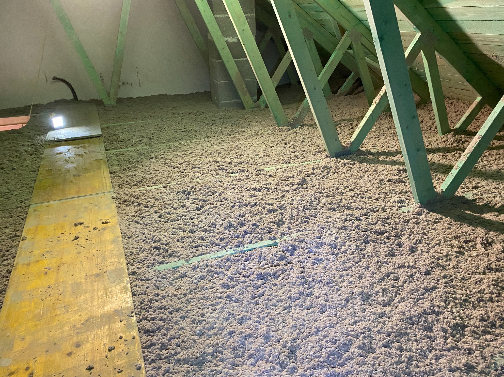 Poddasze z widoczną warstwą izolacji z wełny mineralnej, zielone elementy konstrukcyjne więźby dachowej, deski robocze i oświetlenie punktowe.