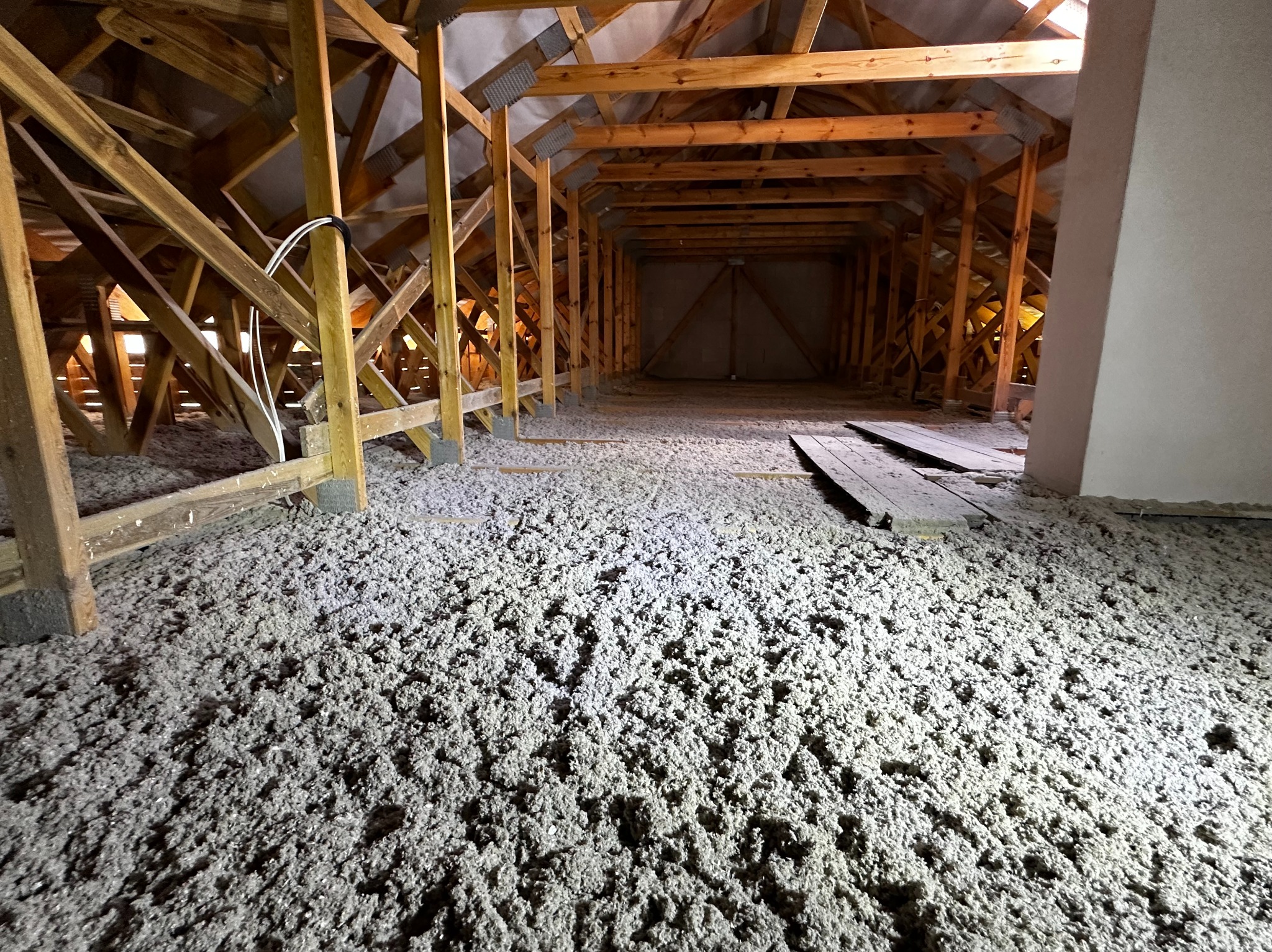Poddasze po ociepleniu wełną mineralną, widoczne drewniane elementy konstrukcyjne dachu i warstwa izolacji. Oświetlenie górne, perspektywa z dołu.