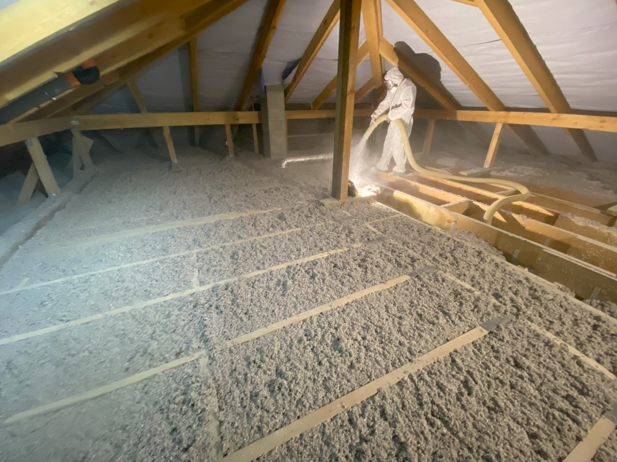 Pracownik w kombinezonie ociepla poddasze domu wełną mineralną, używając specjalistycznego sprzętu. Widoczne drewniane elementy konstrukcji dachu i warstwa izolacji.