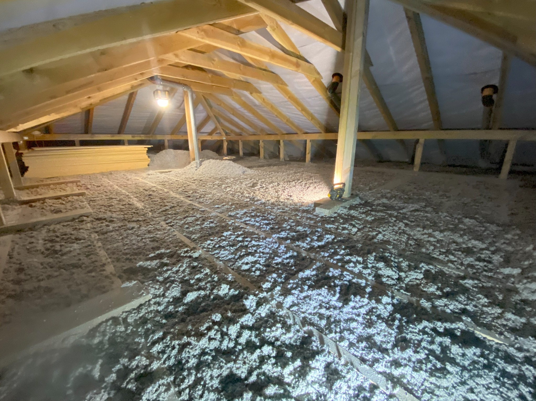 Poddasze z drewnianą konstrukcją i izolacją z wełny mineralnej, oświetlone lampą. Widoczne deski i elementy wentylacyjne. Izolacja równomiernie rozłożona.