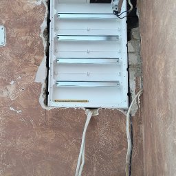 Piotr Wruk Usługi Elektryczne - Biała skrzynka rozdzielcza z szynami DIN, zamontowana w ścianie z dekoracyjnym tynkiem. Widoczne wystające przewody i gruz po kuciu.