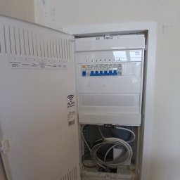 Piotr Wruk Usługi Elektryczne - Otwarta skrzynka elektryczna z bezpiecznikami i przewodami wewnątrz, zamontowana na ścianie. Widoczne oznaczenia obwodów i logo producenta.