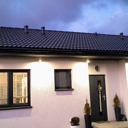 Piotr Wruk Usługi Elektryczne - Elewacja domu jednorodzinnego z zamontowanym oświetleniem zewnętrznym, widoczne lampy nad oknami i drzwiami, czarny dach i okna, wieczorne oświetlenie.