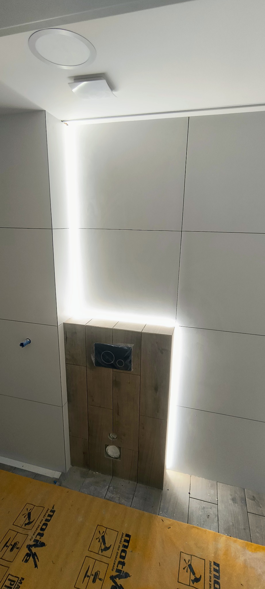 Nowoczesna łazienka w trakcie wykańczania: białe płytki, drewnopodobna zabudowa stelaża WC z podświetleniem LED, widoczne elementy instalacji.