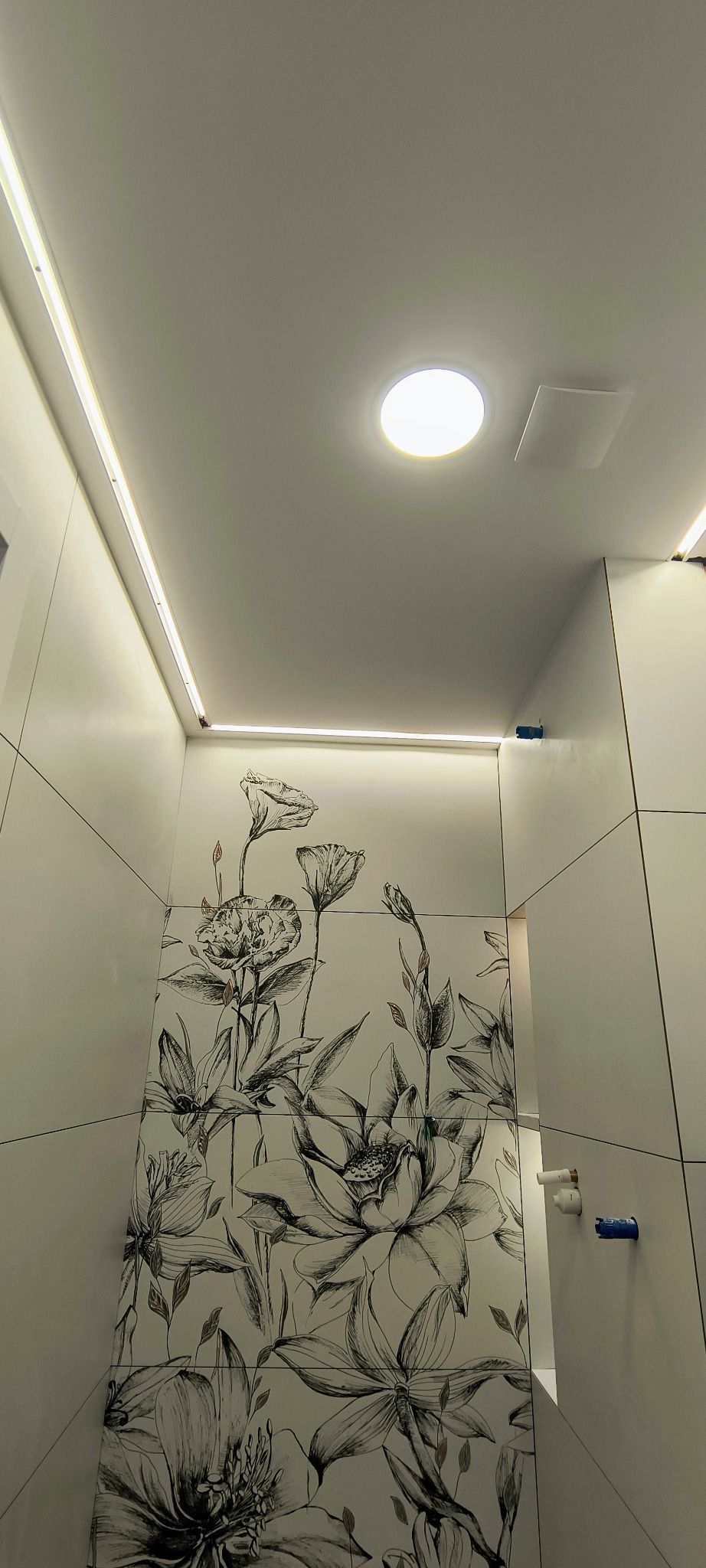 Nowoczesna łazienka z oświetleniem LED podsufitowym i dekoracyjnymi płytkami z motywem kwiatowym. Widok z dołu, podkreślający minimalistyczny design i jasne wykończenie.