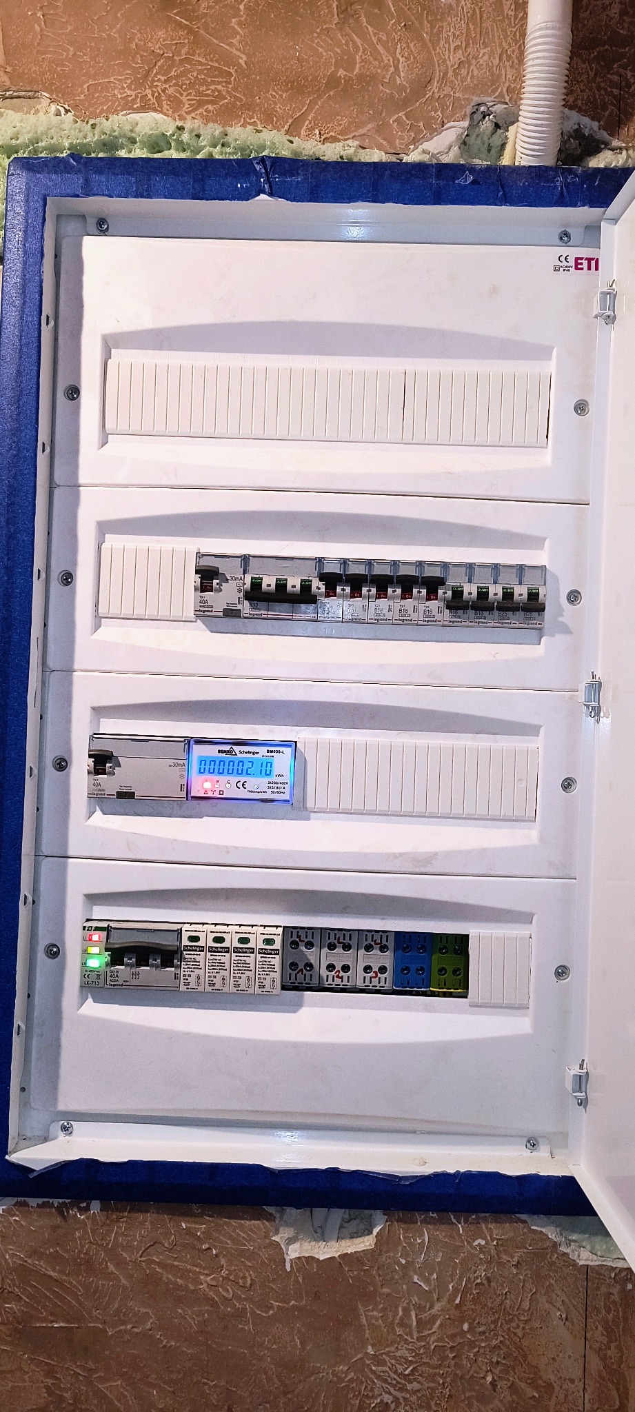 Rozdzielnica elektryczna z zamontowanymi bezpiecznikami, licznikiem energii i zabezpieczeniami przepięciowymi, widok z bliska na ścianie z wykończeniem strukturalnym.