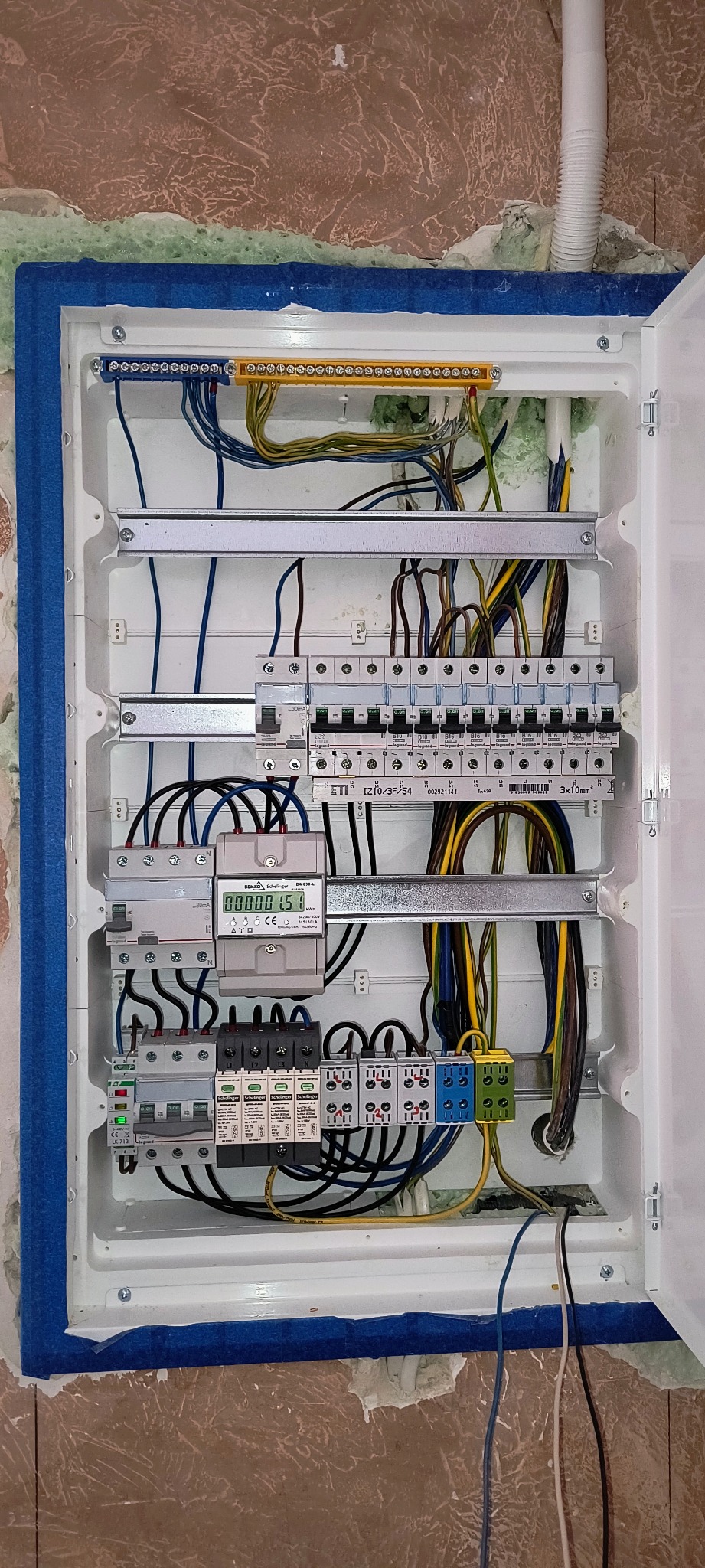 Rozdzielnica elektryczna w trakcie montażu, widoczne kable, zabezpieczenia i licznik. Niebieska taśma malarska zabezpiecza ścianę.