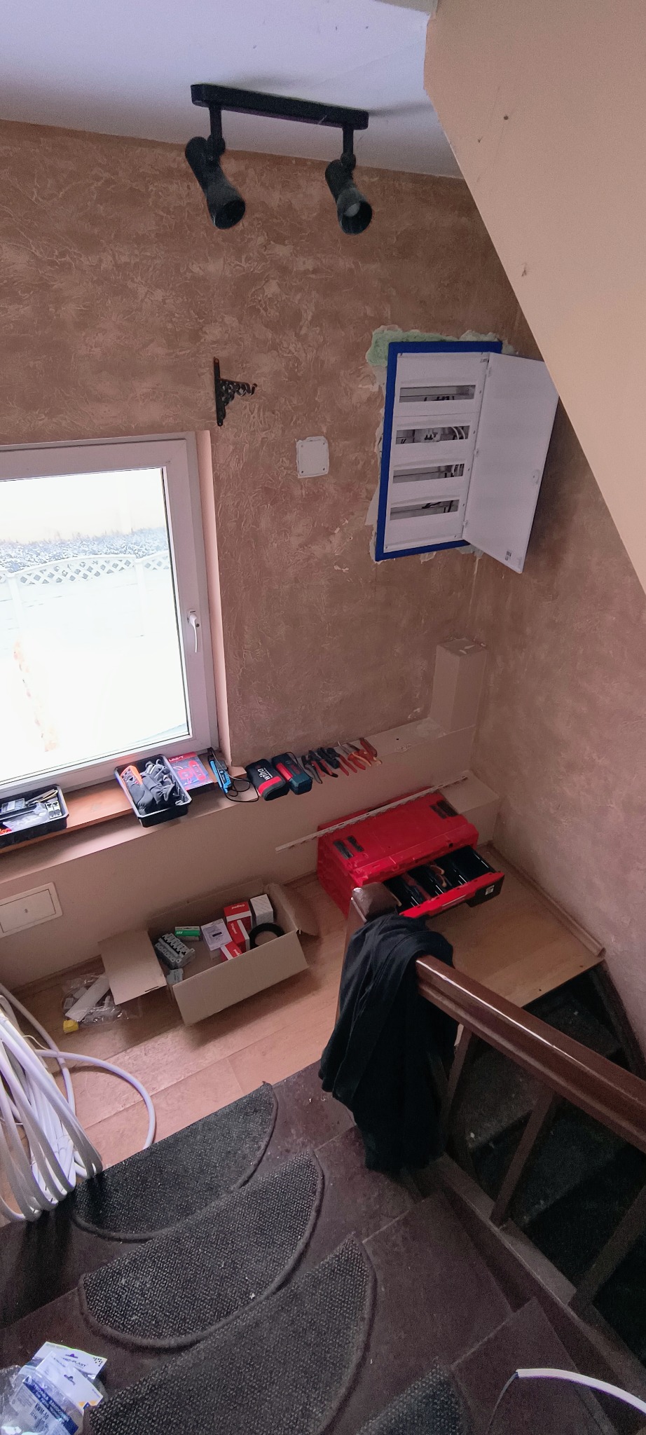 Otwarta skrzynka bezpiecznikowa na ścianie klatki schodowej, narzędzia na parapecie okna, lampa sufitowa. Widok z góry na schody z dywanikami.