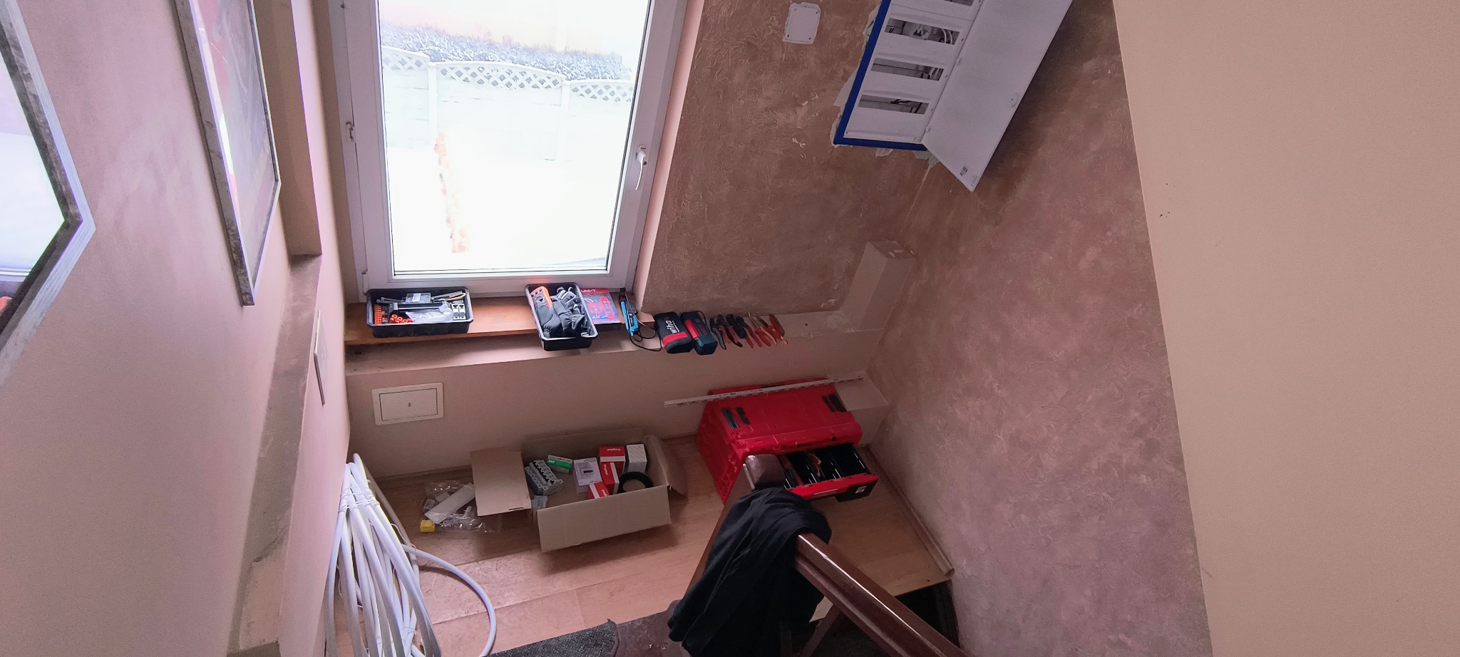 Rozdzielnia elektryczna na ścianie klatki schodowej, skrzynki narzędziowe i akcesoria elektryczne pod oknem dachowym. Widok z góry na narzędzia pracy.