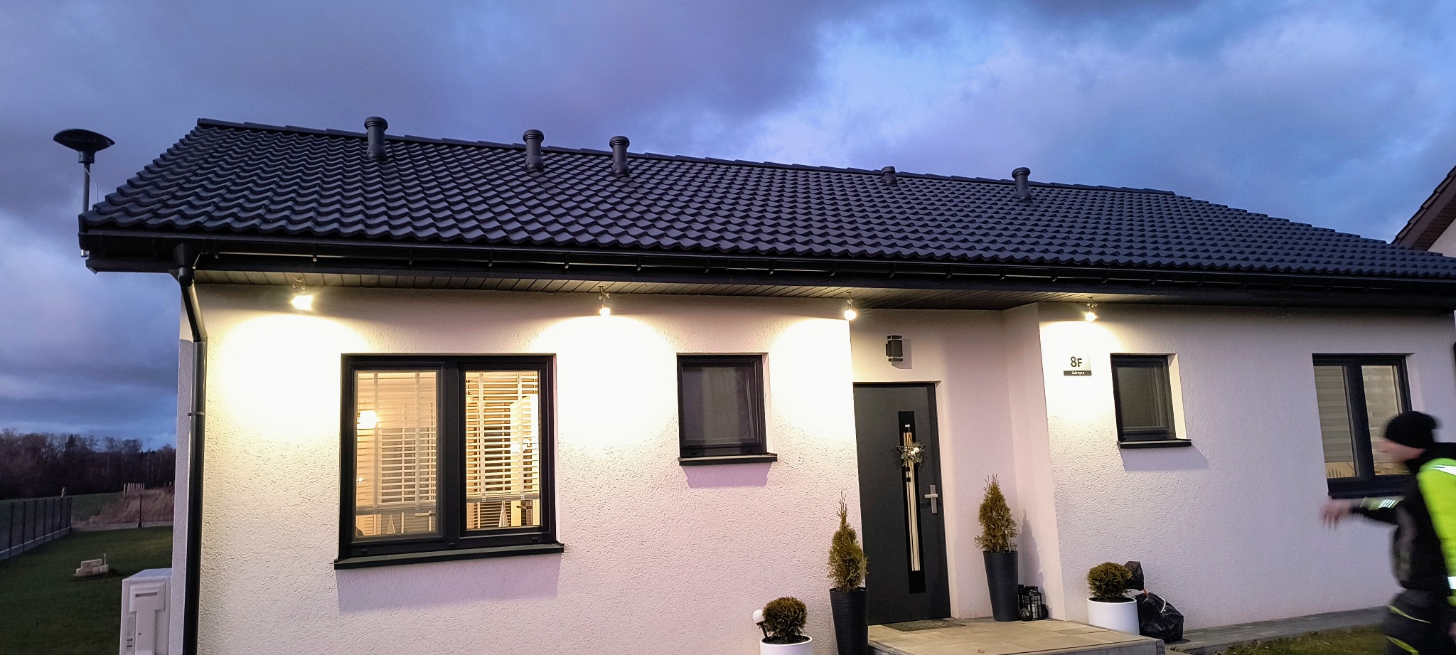 Elewacja domu jednorodzinnego z zamontowanym oświetleniem zewnętrznym, widoczne lampy nad oknami i drzwiami, czarny dach i okna, wieczorne oświetlenie.