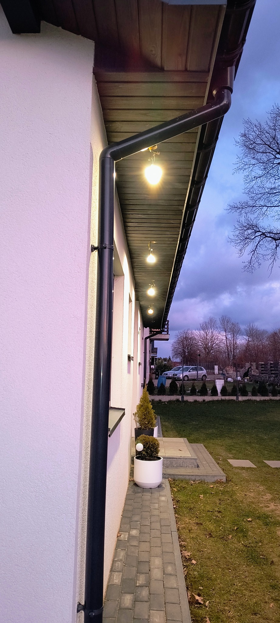 Eleganckie oświetlenie zewnętrzne domu z lampami kulami pod okapem, czarna rynna, biała elewacja i kostka brukowa. Widok na ogród i zachmurzone niebo.