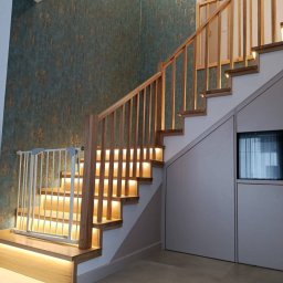 Kamil jonca - Nowoczesne schody z drewnianą balustradą i podświetleniem LED, zabezpieczone bramką dla dziecka. Ściana z wzorzystą tapetą, minimalistyczny design.