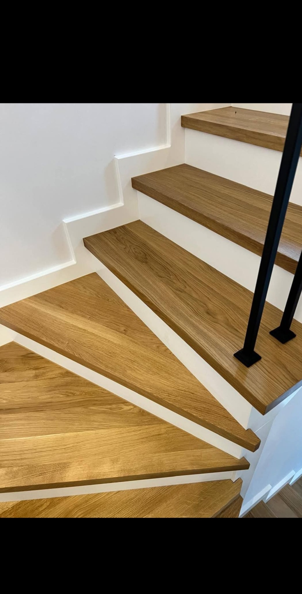 Kątowe ujęcie dębowych schodów z czarną metalową poręczą, białe ściany z dekoracyjnym frezem. Naturalne oświetlenie podkreśla rysunek drewna.