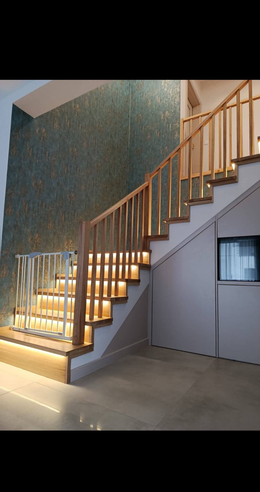 Nowoczesne schody z drewnianą balustradą i podświetleniem LED, zabezpieczone bramką dla dziecka. Ściana z wzorzystą tapetą, minimalistyczny design.