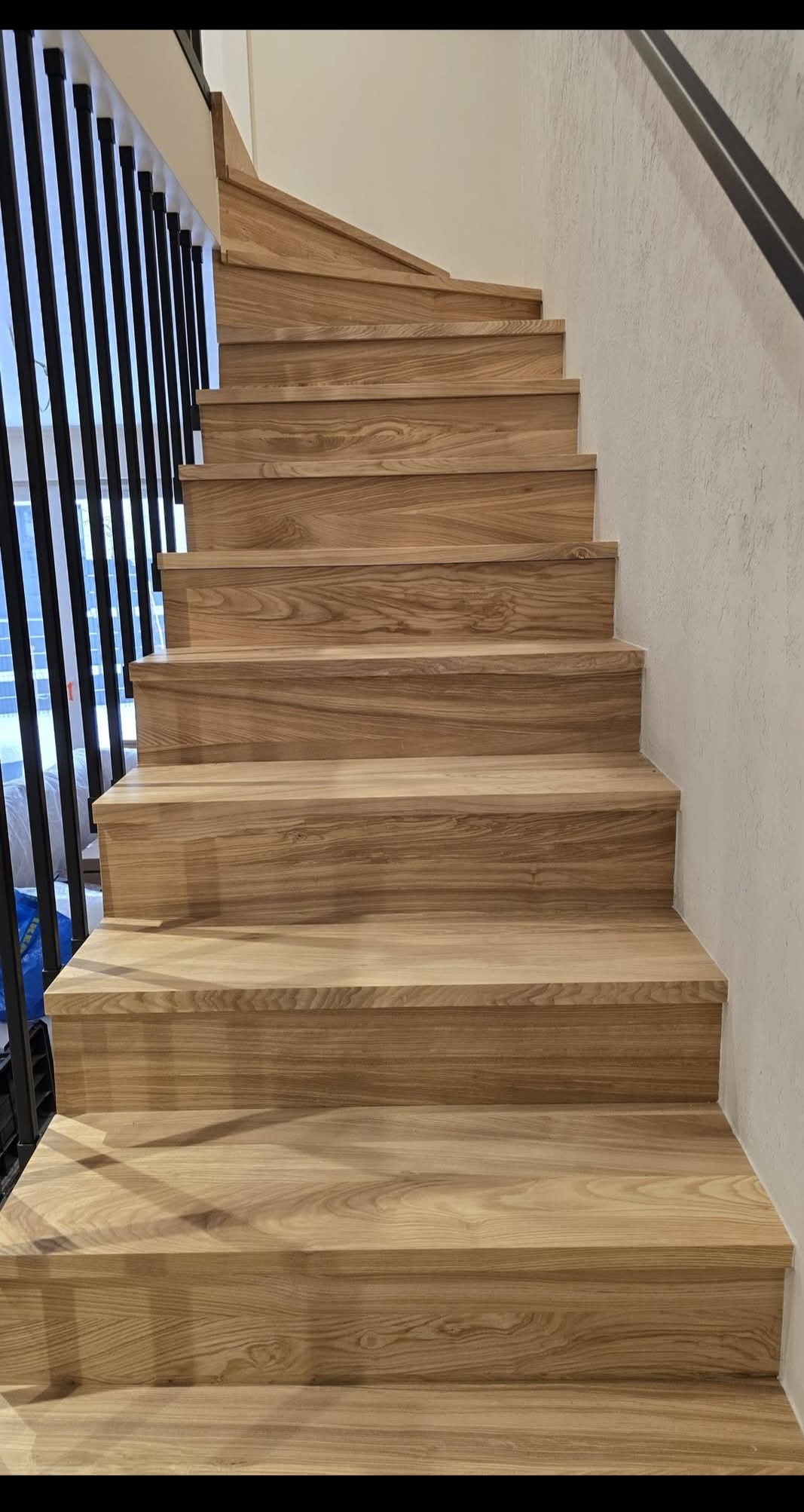 Jasne, drewniane schody z czarną, metalową balustradą, widziane z dołu. Naturalne oświetlenie podkreśla strukturę drewna i minimalistyczny design.