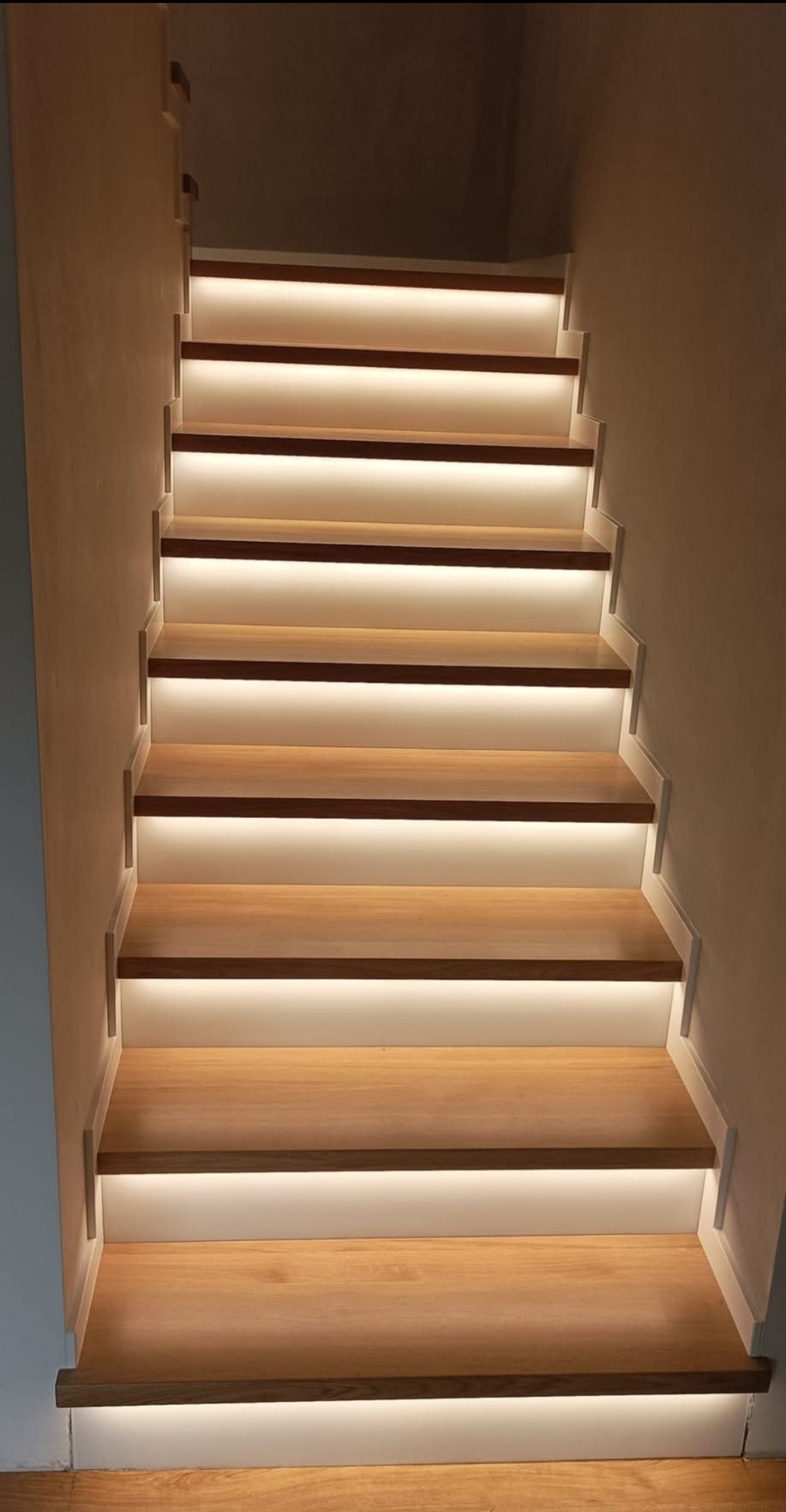 Nowoczesne schody z drewna z subtelnym oświetleniem LED pod każdym stopniem, tworzącym ciepłą atmosferę. Minimalistyczny design, jasne drewno, neutralne tło.