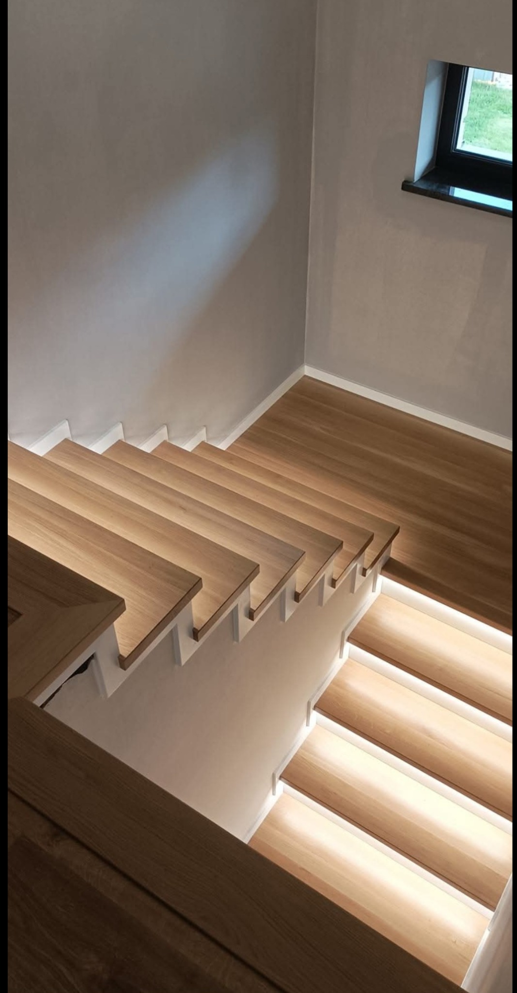 Nowoczesne schody drewniane z podświetleniem LED, widok z góry. Jasne drewno kontrastuje z białymi elementami konstrukcyjnymi i szarą ścianą. Minimalistyczny design.